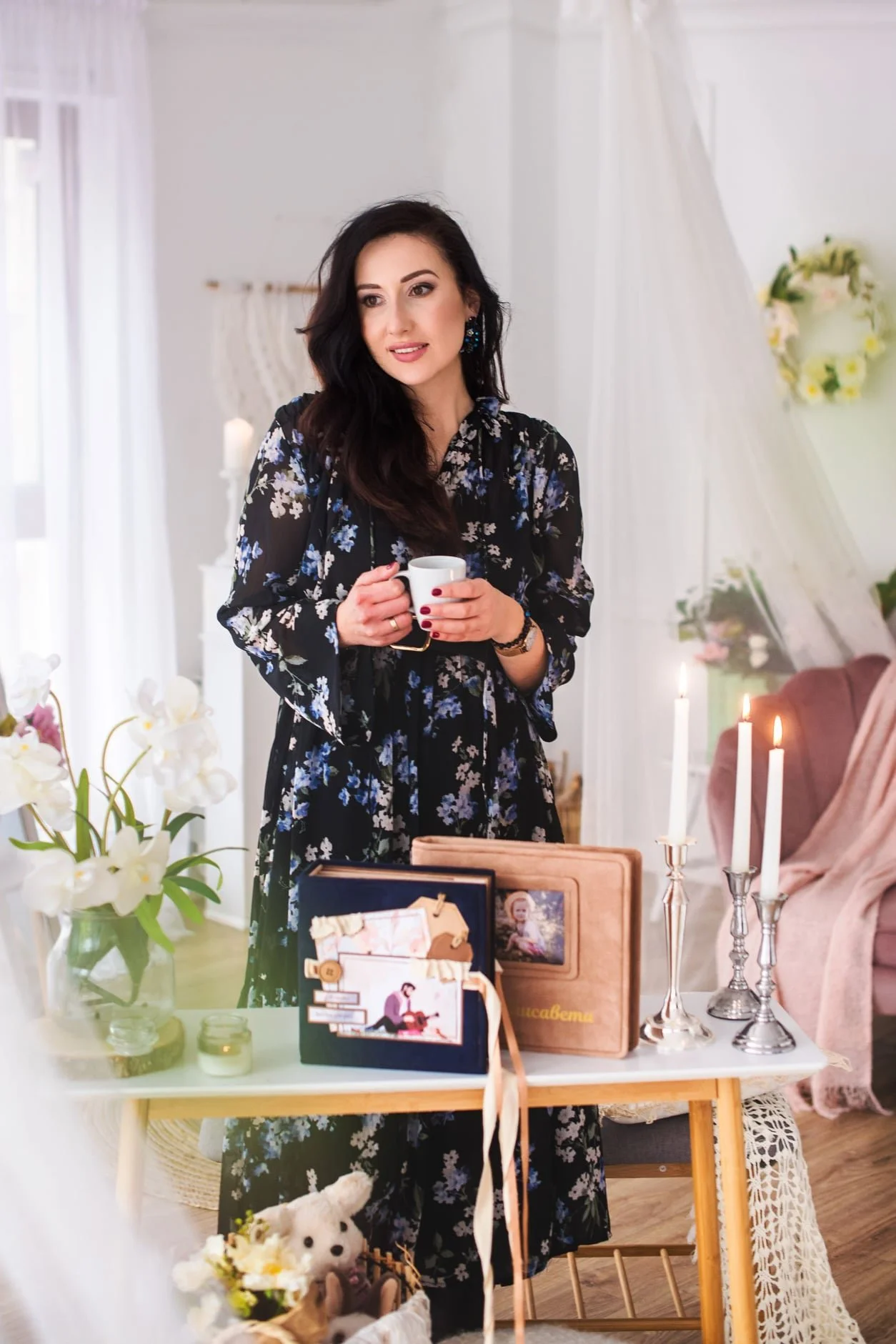 Жена в тъмна рокля на цветя пред маса с ръчно направени албуми, свещи и вази с цветя