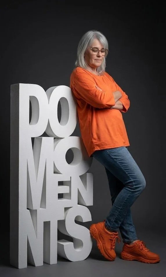 Happy DO with gray hair and glasses wearing an orange sweatshirt, blue jeans, and orange sneakers, leaning against large 3D letters spelling "DO MOMENTS" on a dark gray background.