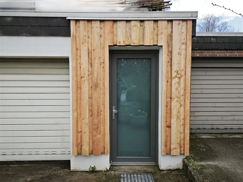 Modernisierter Garagennebeneingang mit einer vertikalen Boden-Deckel-Schalung aus naturbelassenem Holzfassaden-Profil, eingefasst zwischen zwei Garagentoren.