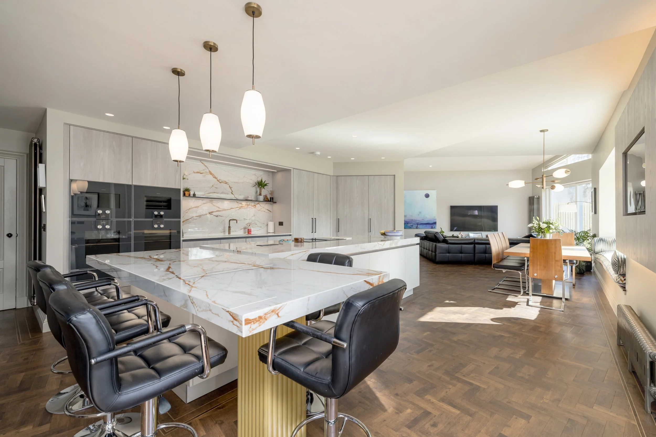Open-concept kitchen and living room with marble island, black bar stools, and modern decor, featuring a dining table with chairs, a sofa, and a flat-screen TV.
