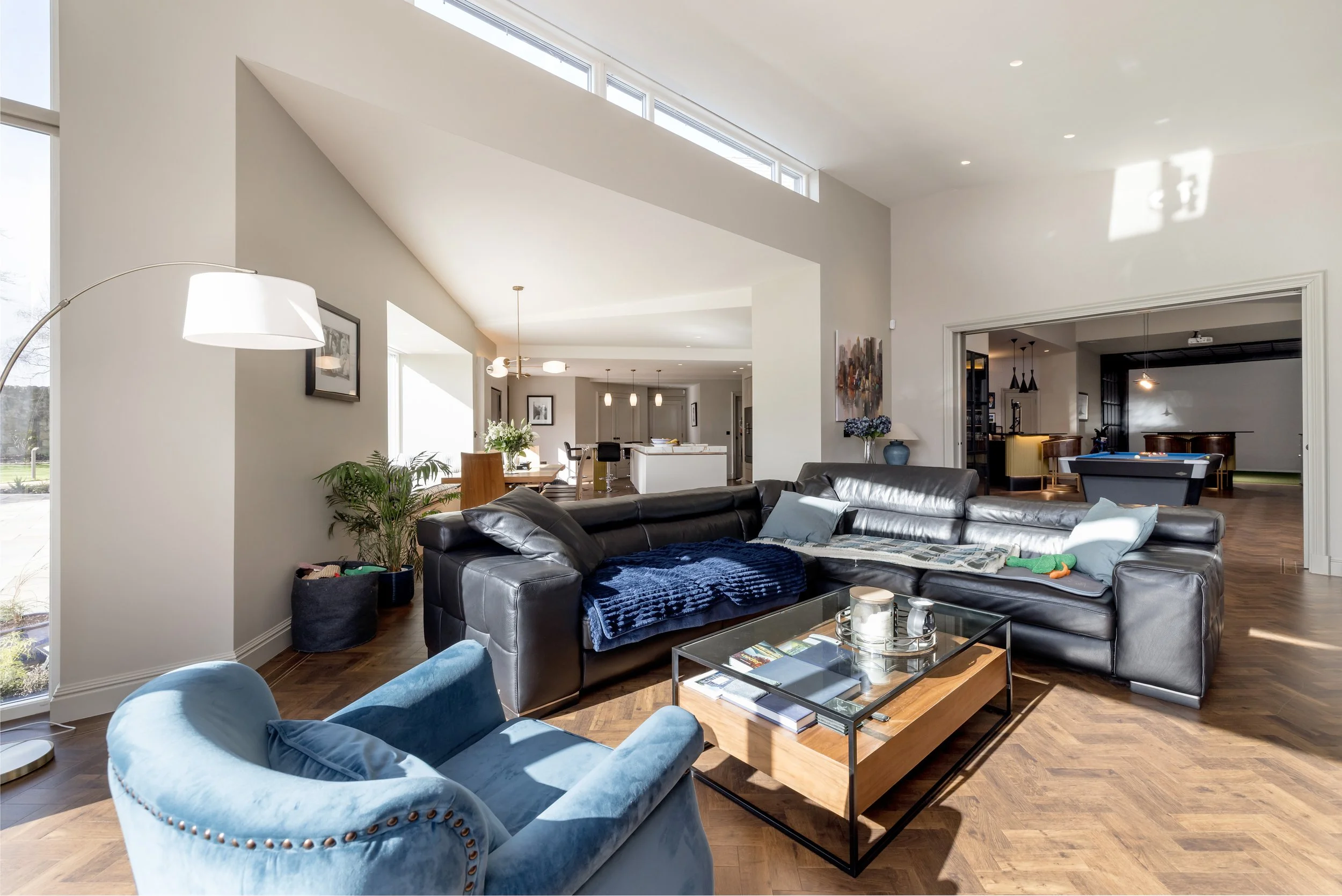 Living room with black leather sectional sofa, blue velvet armchair, glass and wood coffee table, and sunlight coming through large windows.
