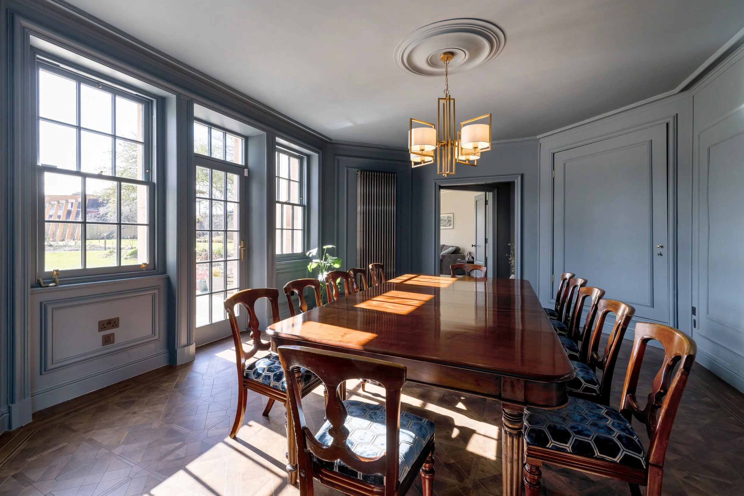 A formal dining room with a polished wooden table and twelve matching chairs, large windows with sunlight streaming in, light blue walls with decorative molding, and an elegant ceiling light fixture.