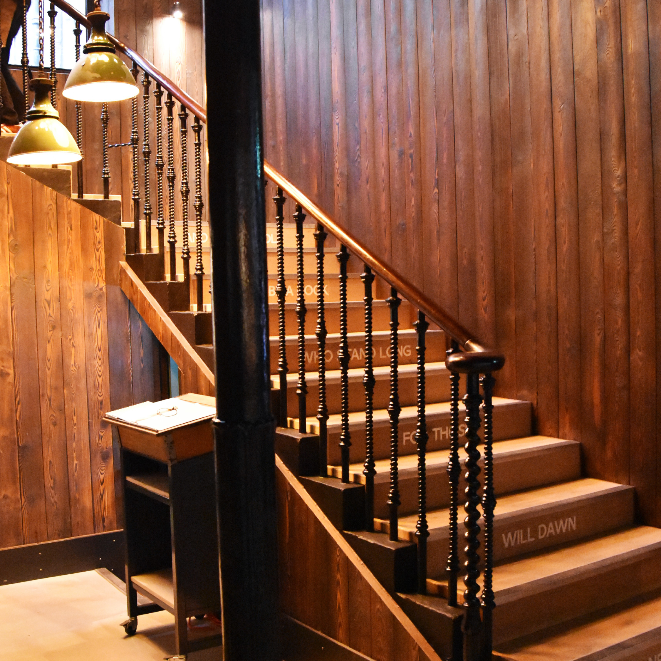 Wooden staircase with black spindles and wooden handrail, with motivational quotes written on the steps, inside a warmly lit room with wooden walls and lamps.