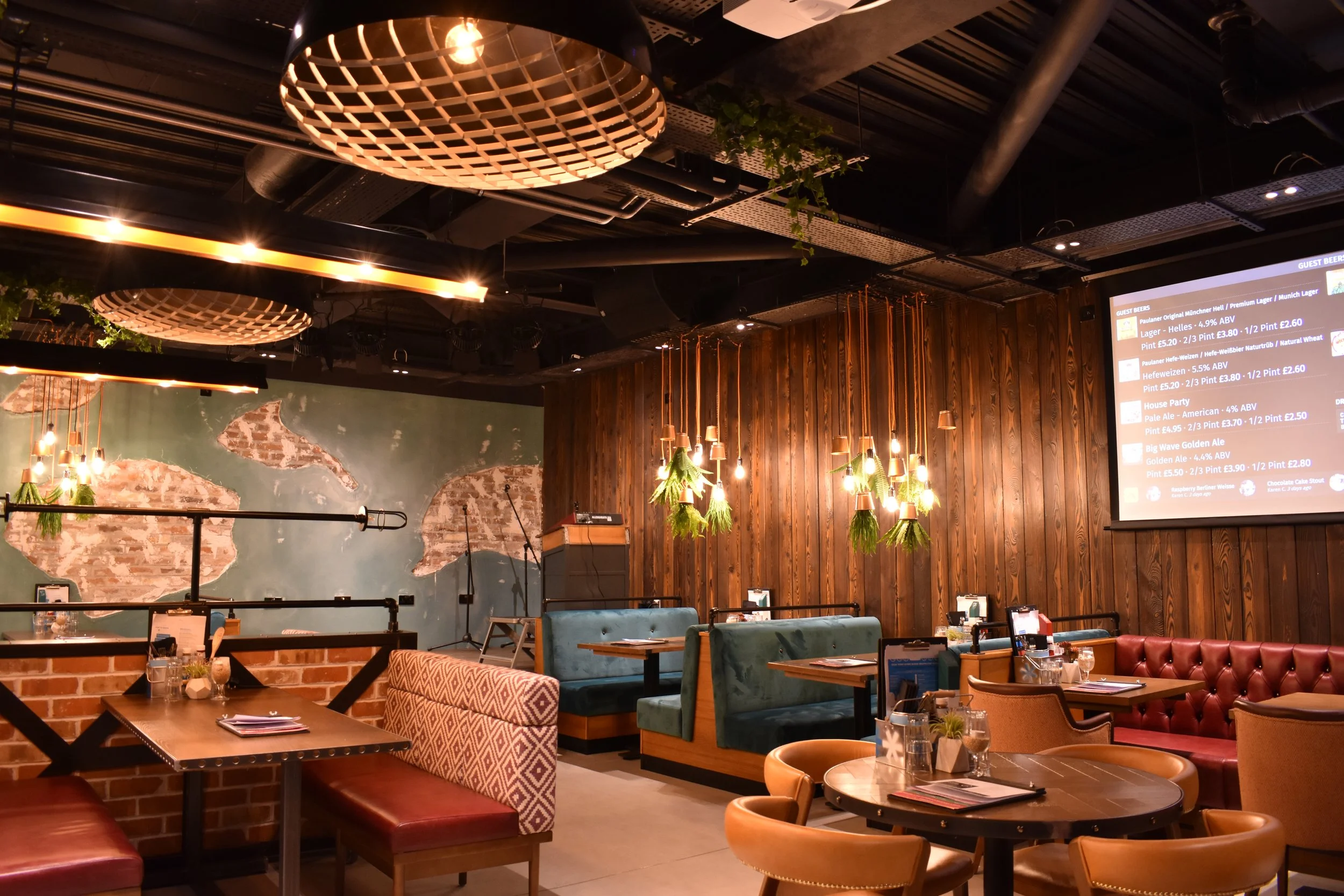 Interior of a cozy restaurant or pub with wooden walls, hanging pendant lights with green plant decorations, a large screen displaying the menu, and various tables and chairs for dining.