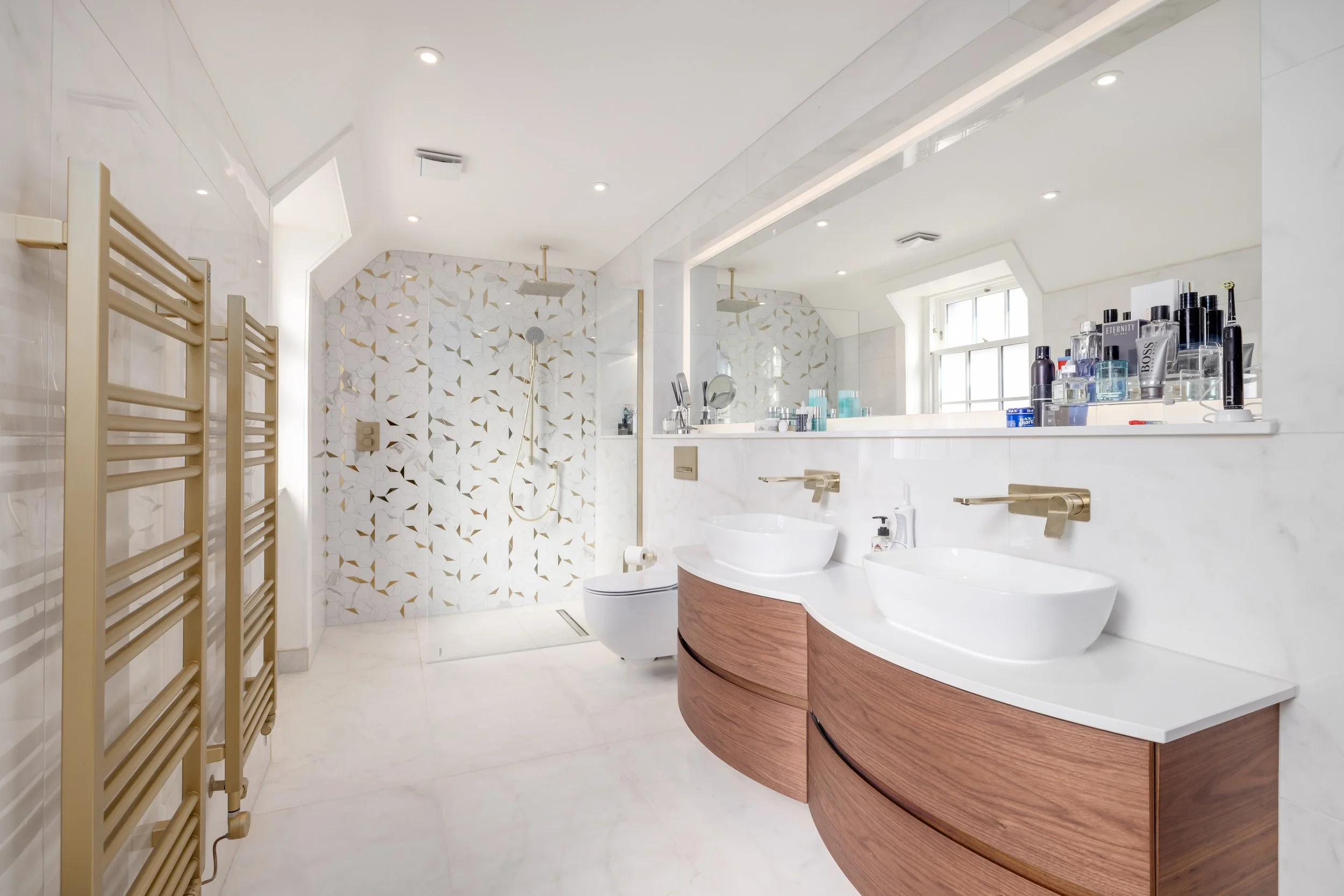 Modern bathroom with double sinks, wall-mounted faucets, large mirror, shower with rain head and patterned tile wall, wooden cabinetry, towel radiator, and toiletries.