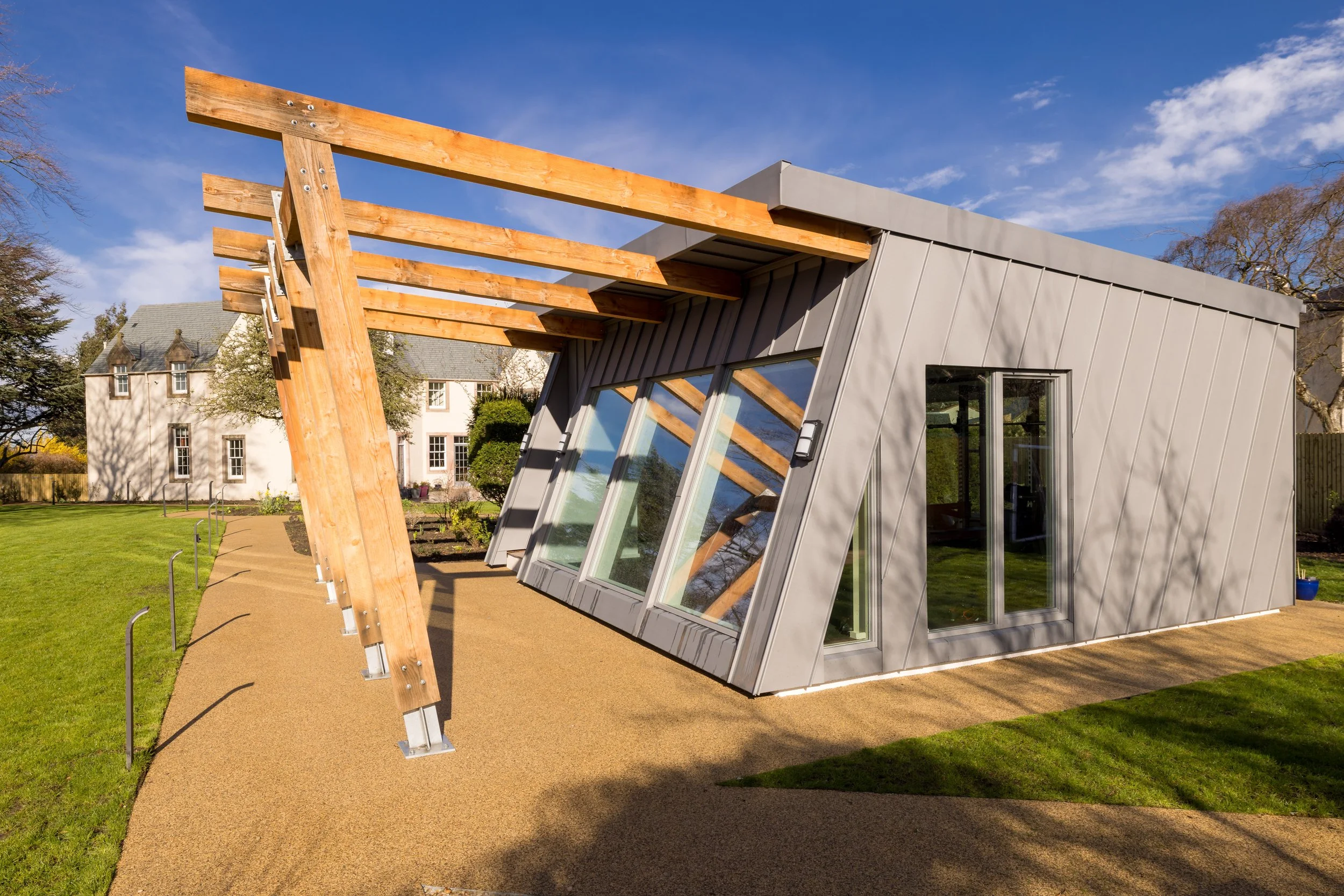 Modern building with angled walls, large windows, and wooden beams extending from the roof, set in a landscaped yard with a grass lawn and pathway.
