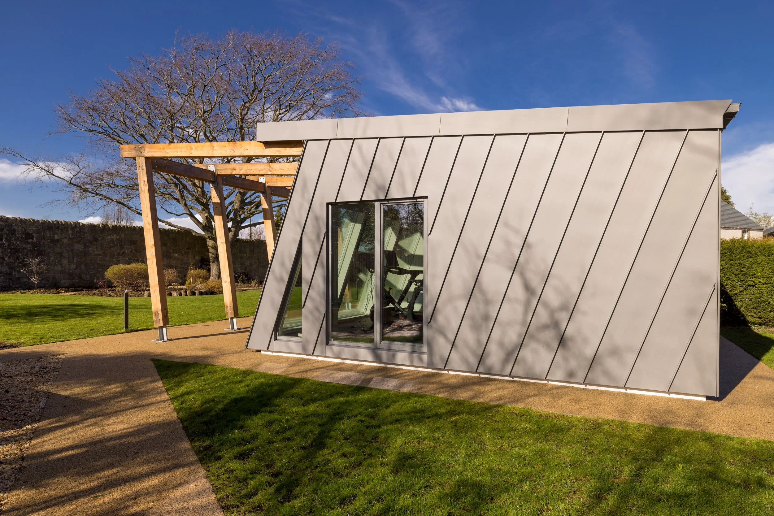 Modern building with slanted gray metal exterior, glass sliding door, and wooden pergola structure in outdoor garden area with green grass, trees, and clear blue sky.