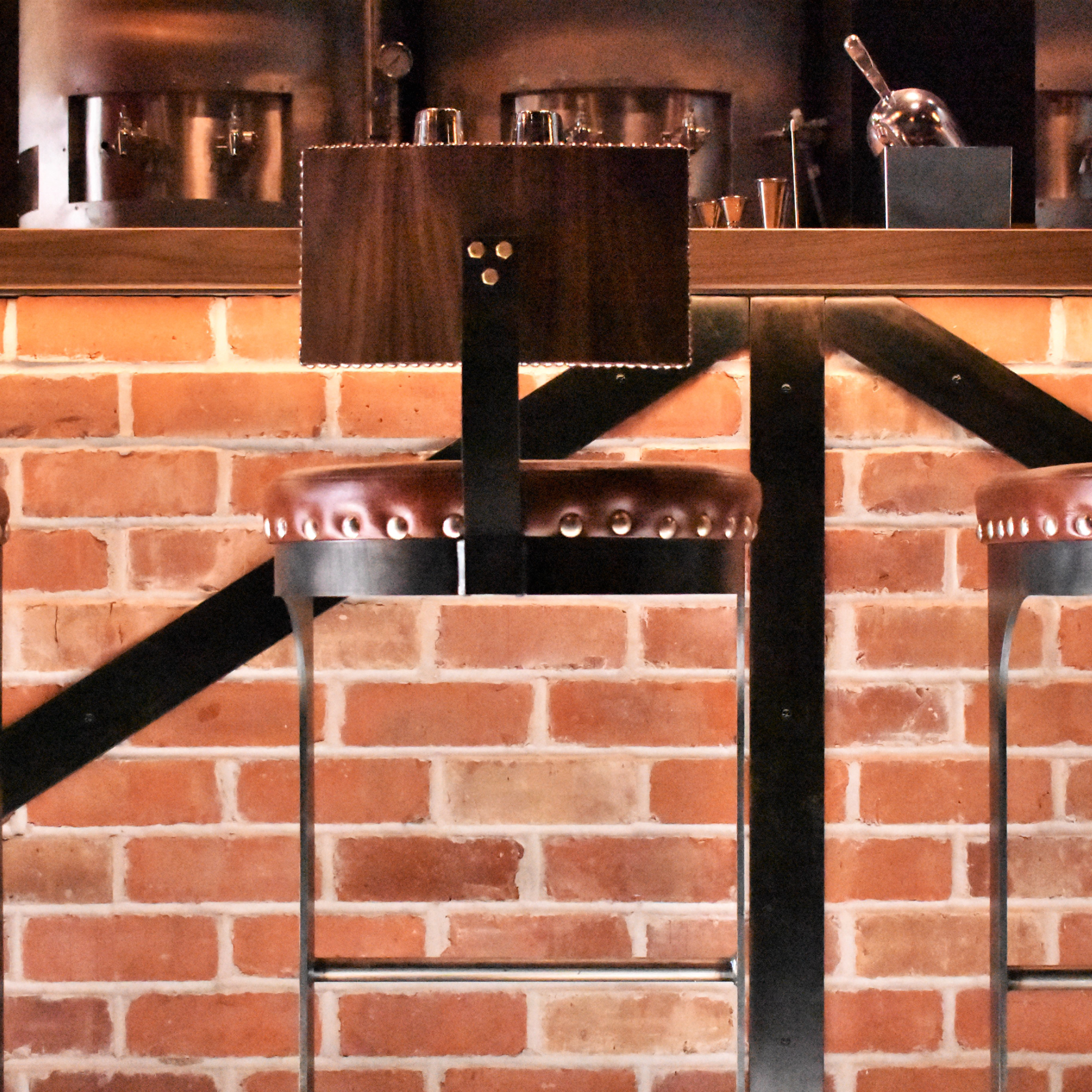 Bar stool with a brown leather seat and metal legs, in front of a brick wall in a restaurant or bar interior. A dark wood table and bar equipment are visible in the background.