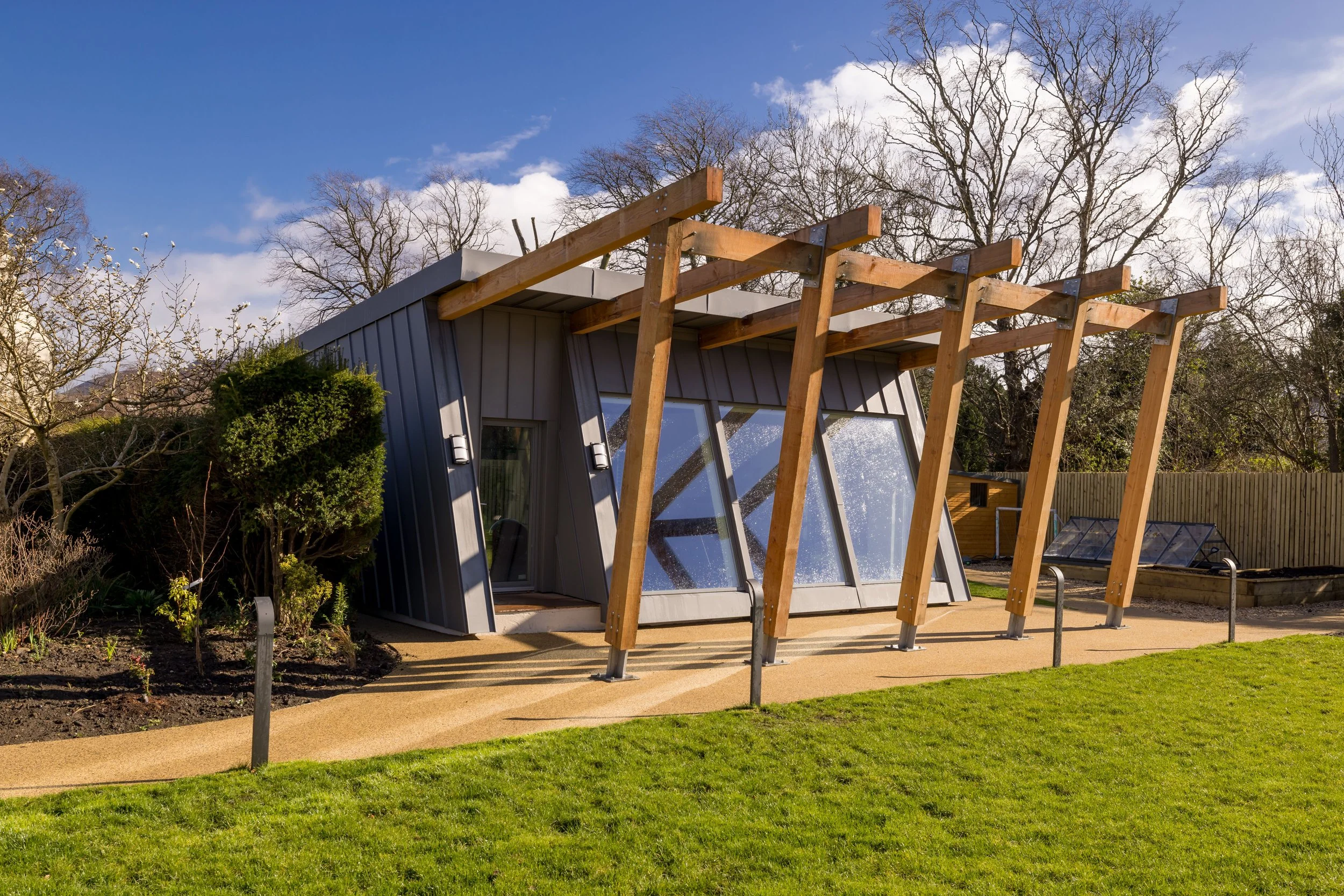 Modern house with angled metal exterior and large glass windows, surrounded by grassy yard, trees, and garden beds, under a partly cloudy sky.