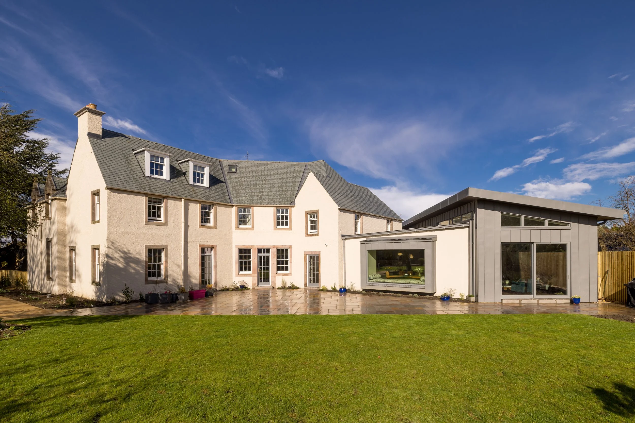 A large house with white stucco walls and grey roof, and a modern extension with large windows, a patio, and a well-maintained green lawn under a blue sky.