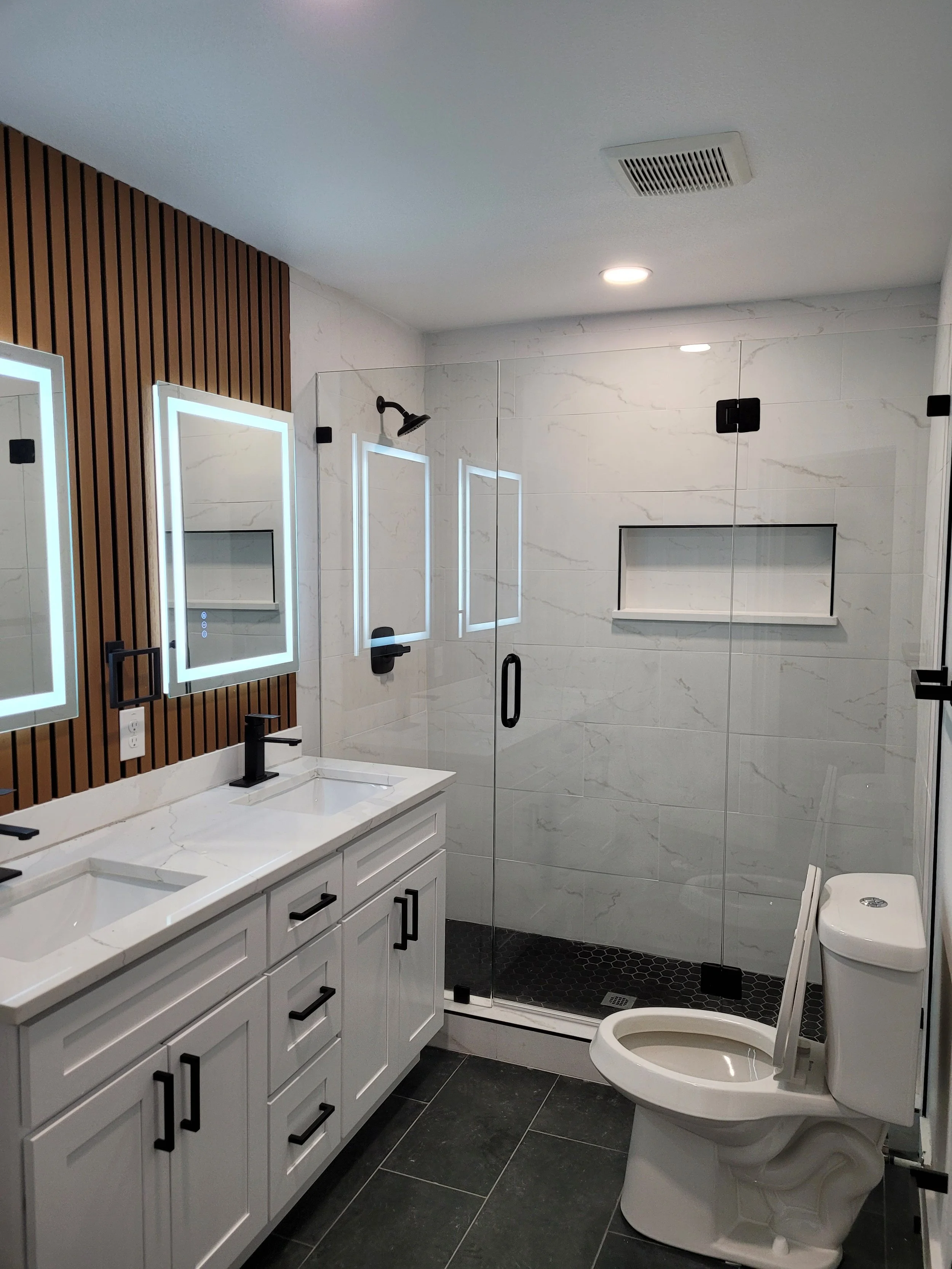 Modern bathroom with double vanity, illuminated mirrors, walk-in shower with glass door, and toilet, featuring white marble walls and black fixtures.
