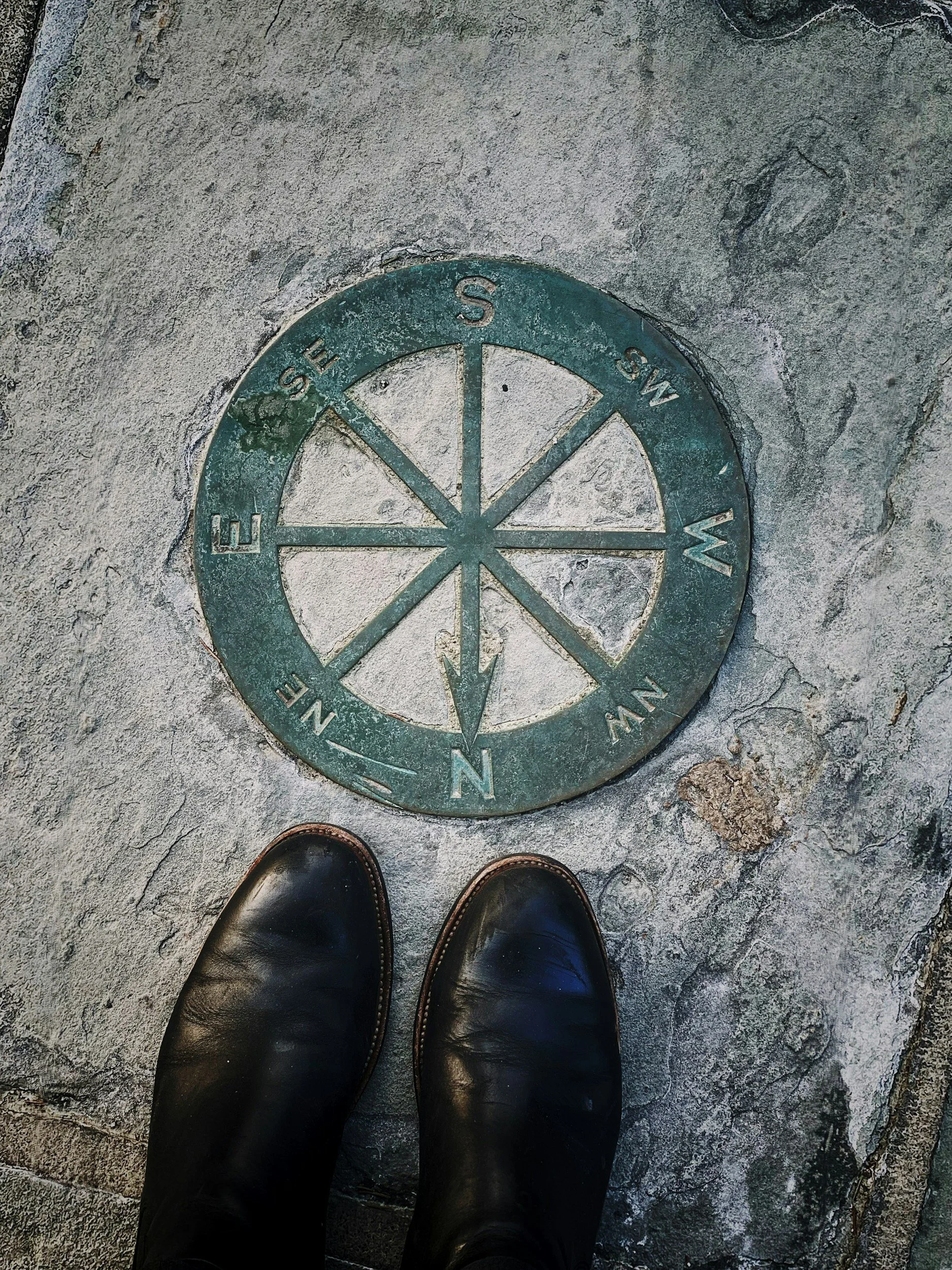 Ground marking of a compass rose indicating directions North, South, East, and West, with a person's black leather shoes visible at the bottom of the image.