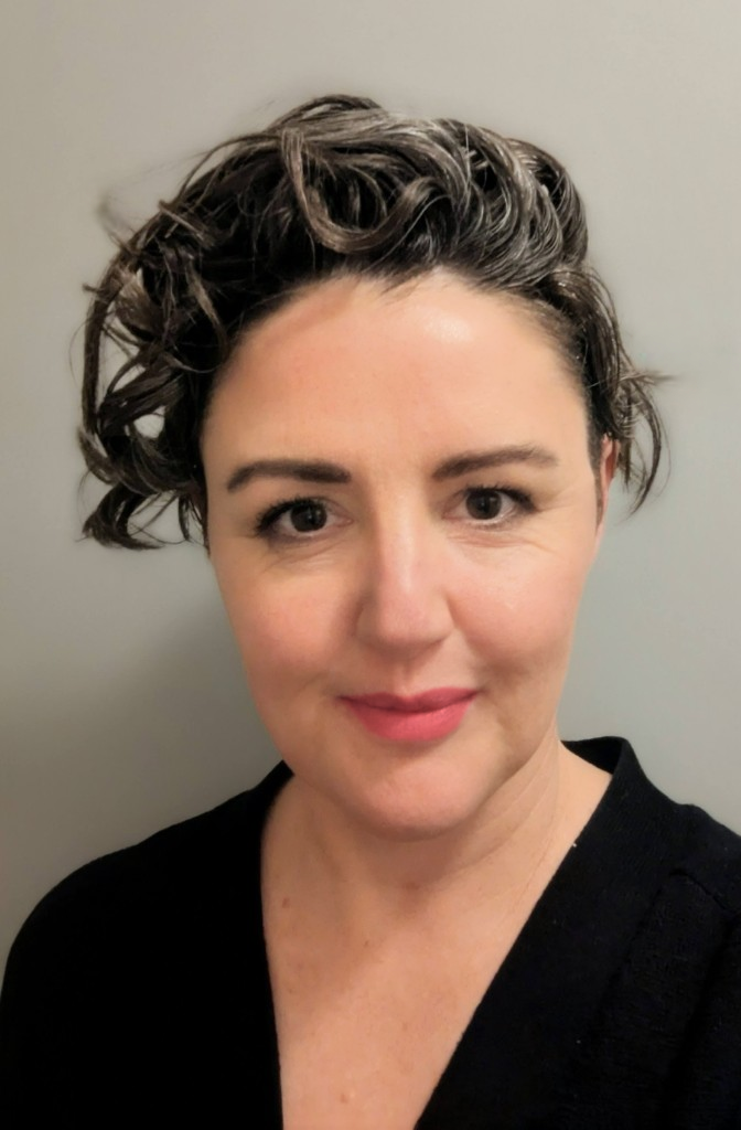 A woman with short, curly brown hair and fair skin taking a selfie against a plain gray background, wearing a black top.