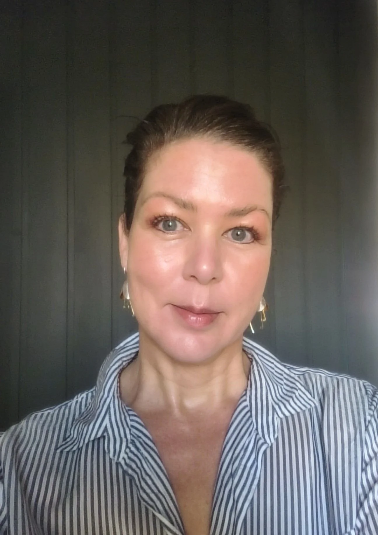 A woman with brown hair in an updo, wearing a striped shirt and earrings, standing in front of a dark wooden wall, looking at the camera.
