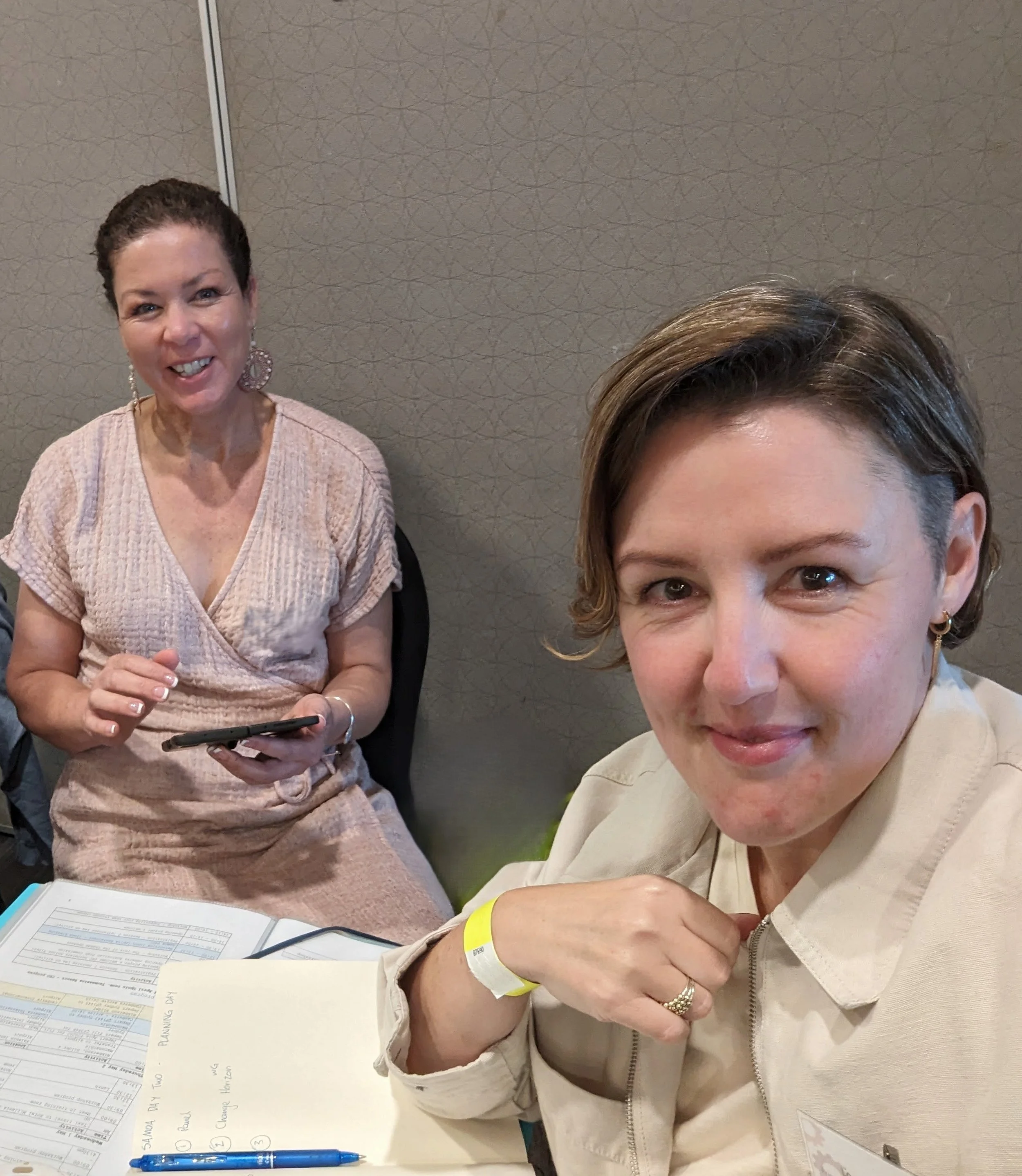 Two women sitting at a table, smiling at the camera. One woman is holding a phone, the other has a notepad and pen, with papers and a yellow wristband visible.