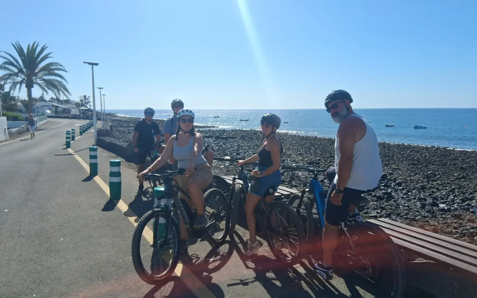 Gruppe von fünf Menschen mit Fahrrädern am Meer, im Sonnenschein, auf einer promenade, mit Palmen im Hintergrund.
