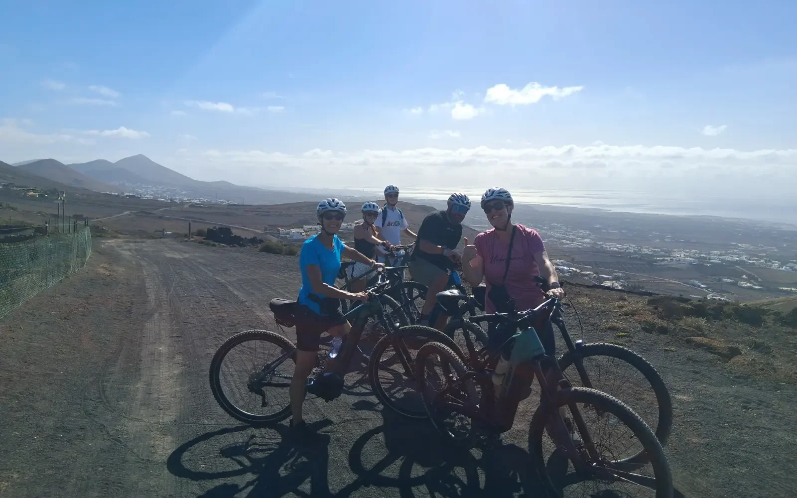 Fünf Fahrradfahrer mit Schutzhelmen auf einer Hügelstraße mit Blick auf das Meer und Berge im Hintergrund.