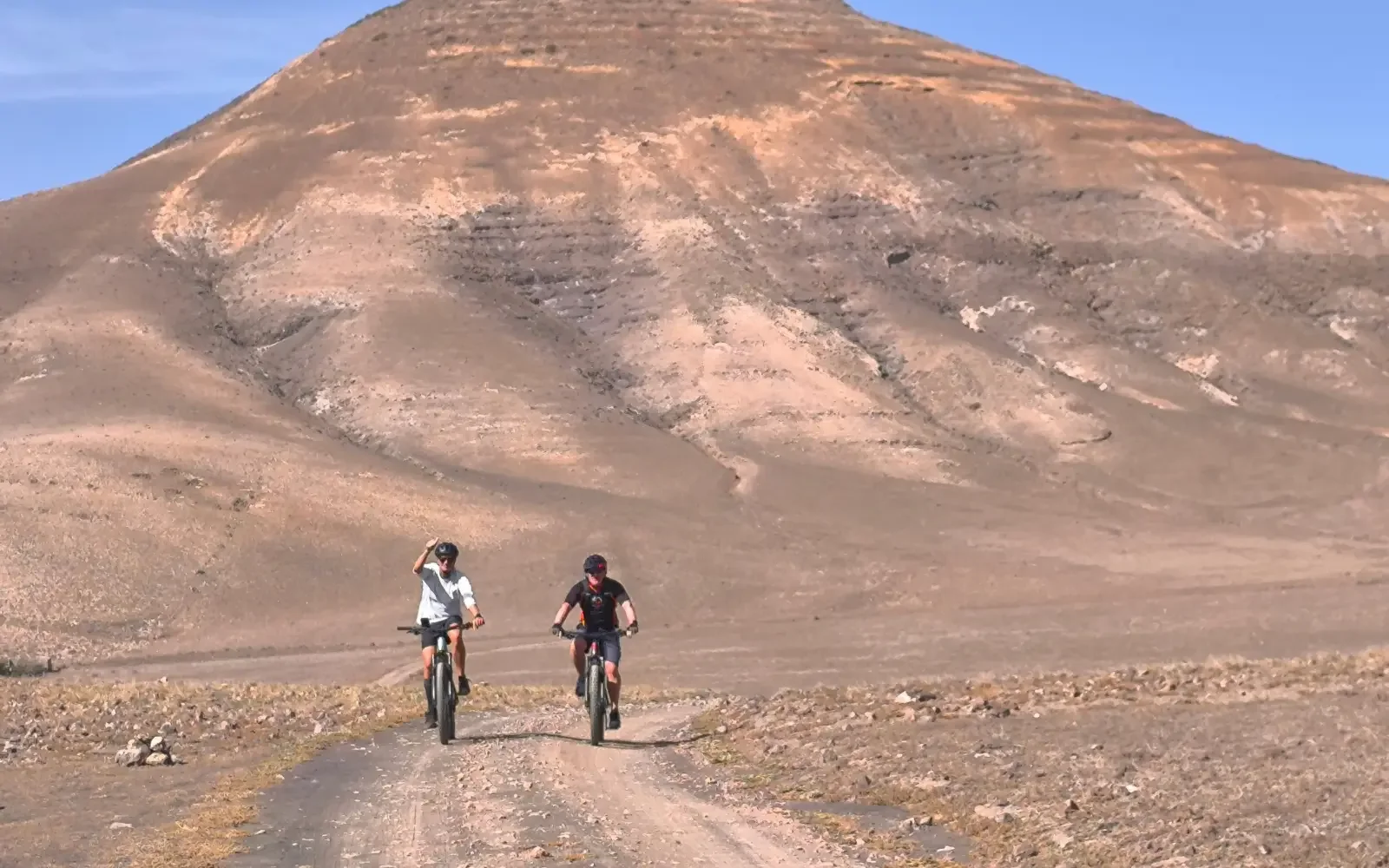 Zwei Radfahrer auf einer staubigen Straße in einer kargen, bergigen Landschaft.