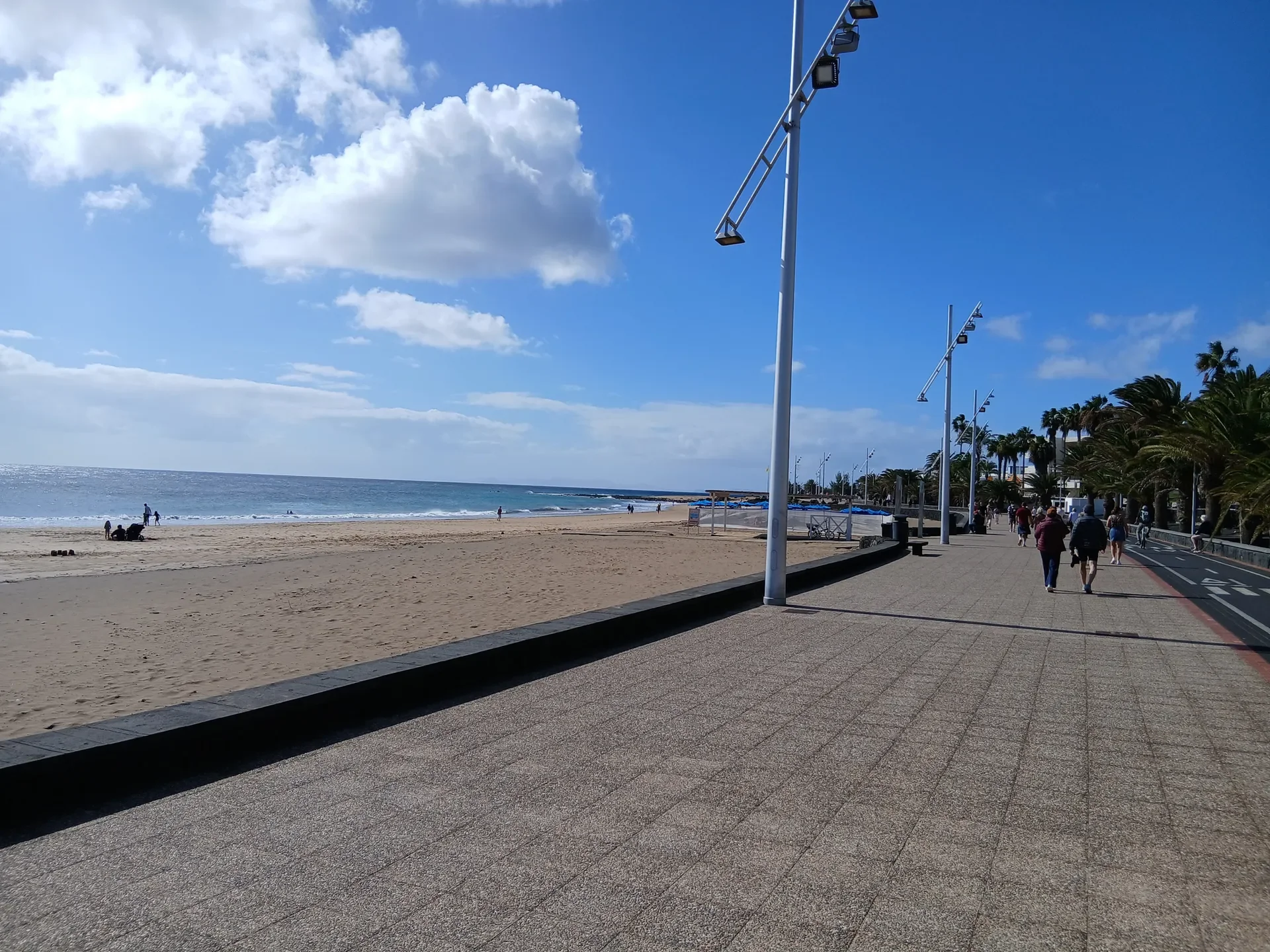 Strandpromenade mit Menschen, Palmen, Leuchttürmen, Blick aufs Meer und Wolken am Himmel.