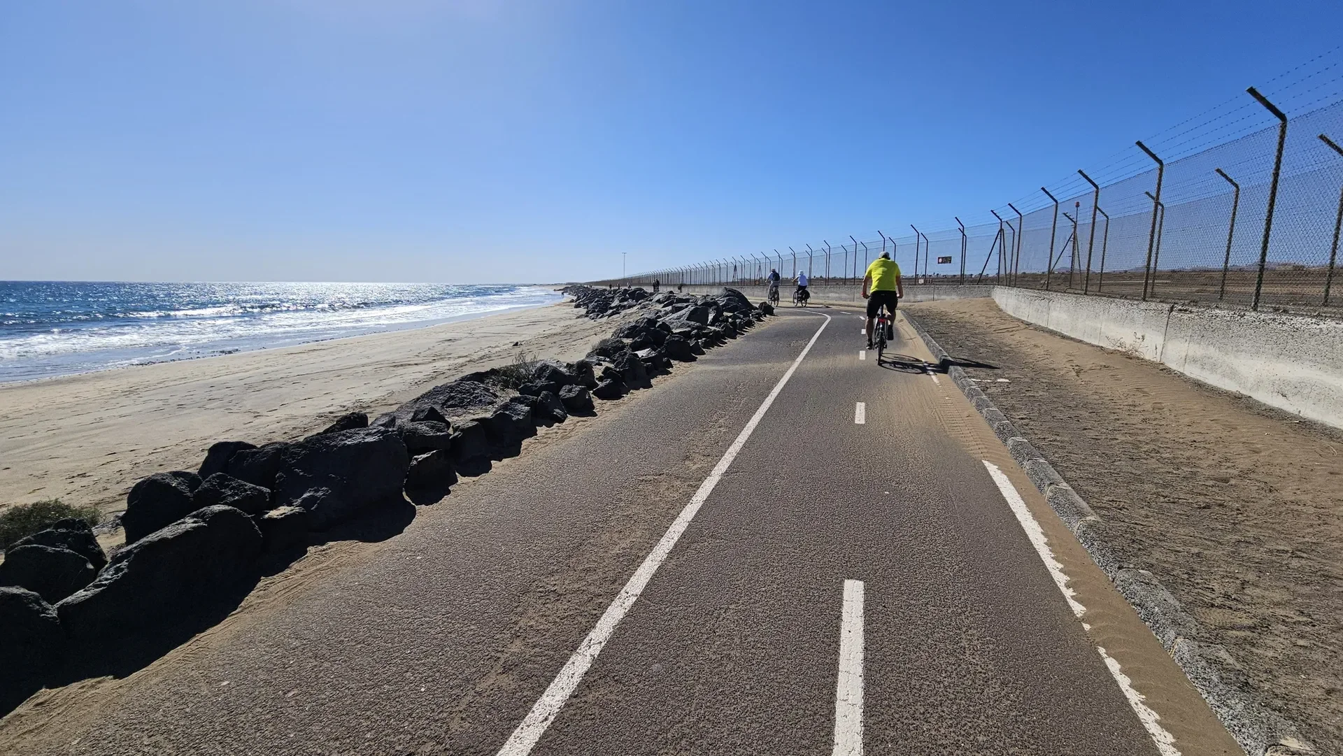 Radfahrer, die auf einer asphaltierten Fahrradbahn am Strand entlangfahren, mit Meer und Sand im Hintergrund, unter klarem blauen Himmel.