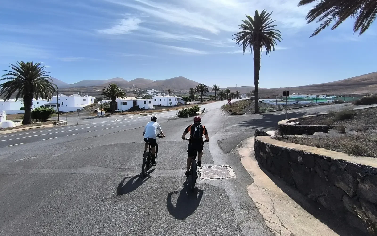 Zwei Radfahrer fahren auf einer Straße in einer sonnigen, trockenen, ländlichen Gegend mit weißen Gebäuden und Palmen im Hintergrund, Berge im Horizont.