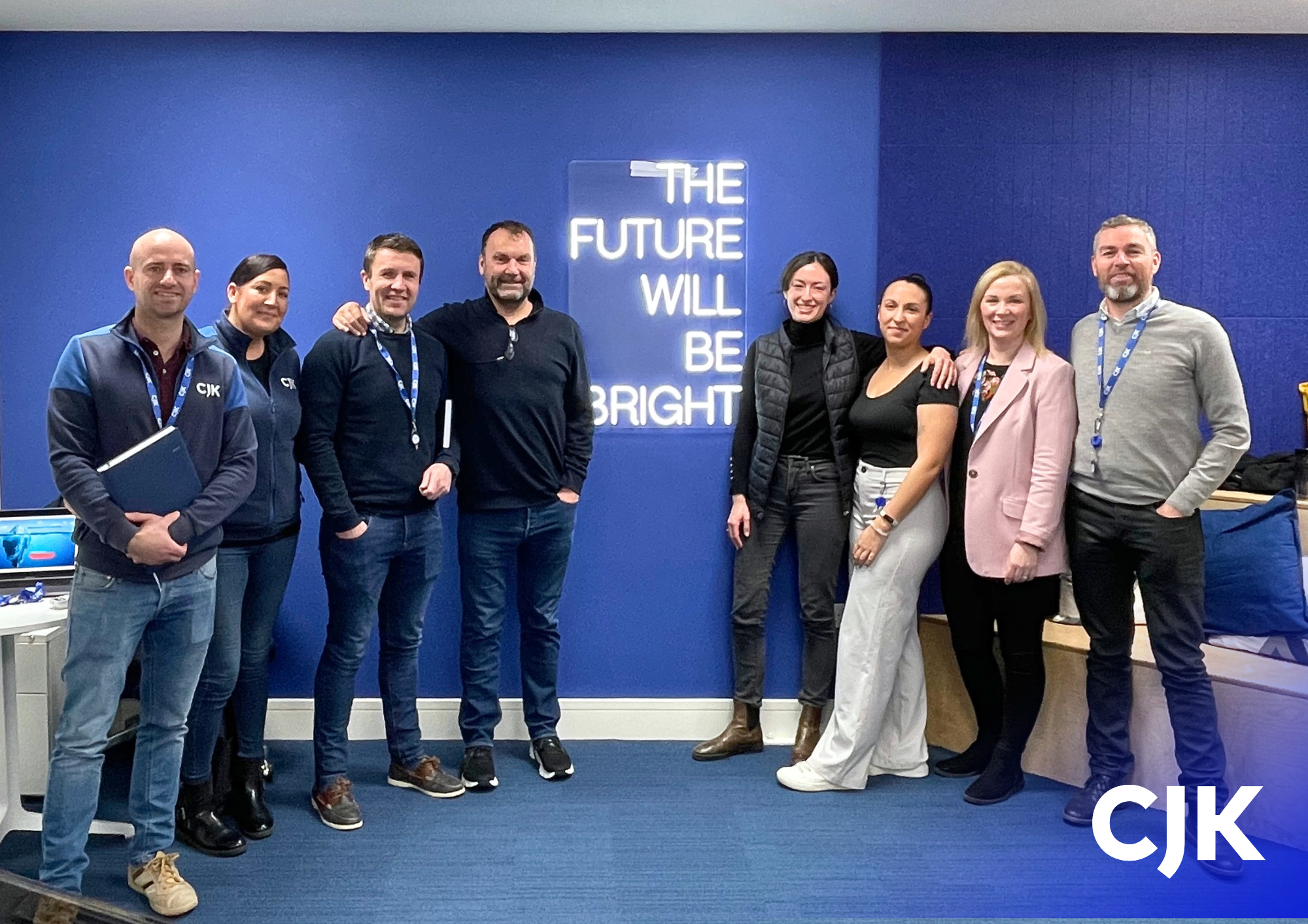 Group of the CJK sustainability committee standing together, smiling, with a neon sign that reads 'The future will be bright' on a blue wall behind them.