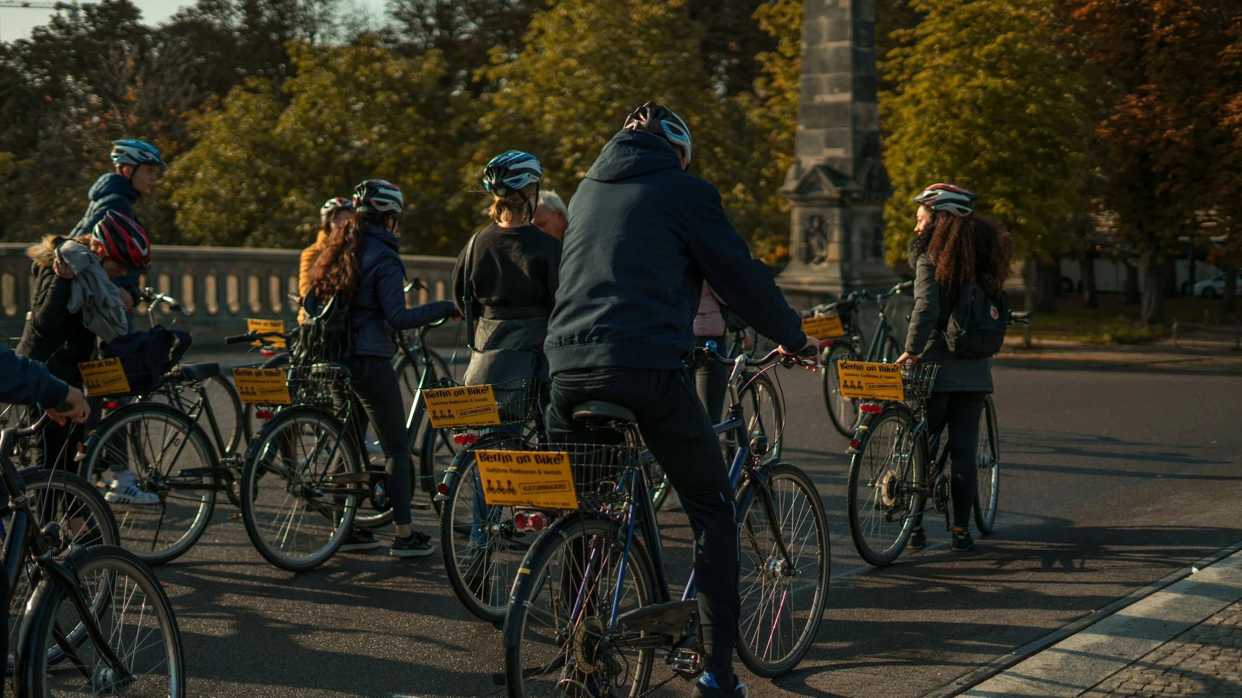 Group of people wearing helmets standing with their bicycles on a city street, with a monument and trees in the background.