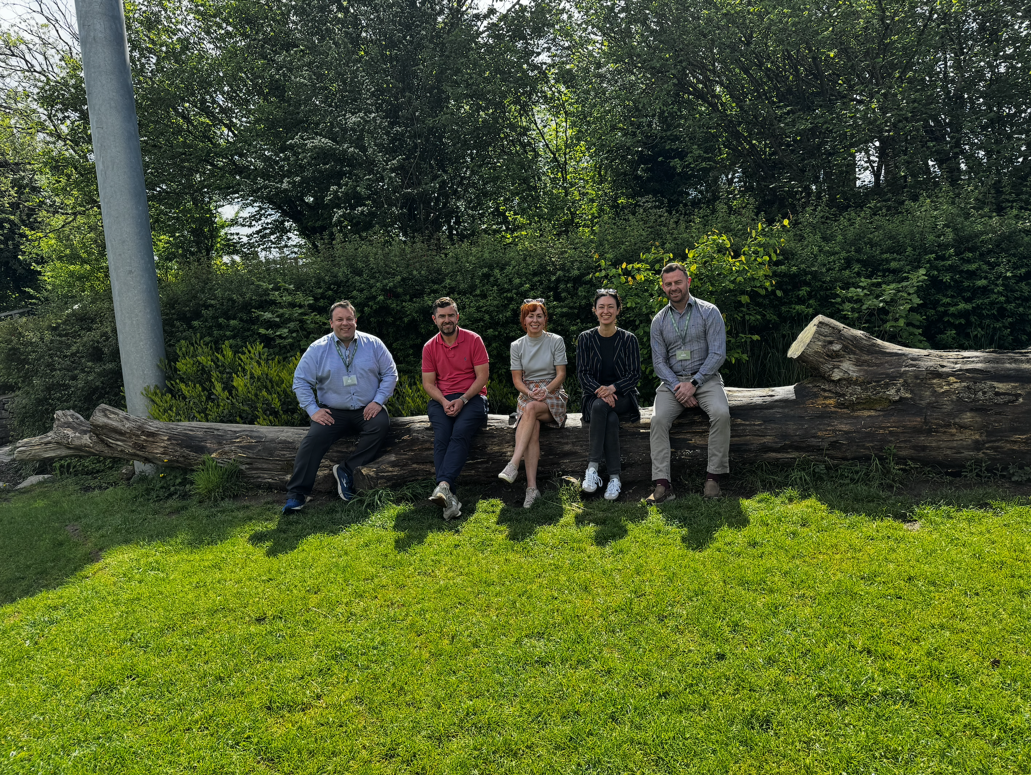 Five people sitting on a fallen log outdoors on a sunny day, with lush green trees and bushes behind them.