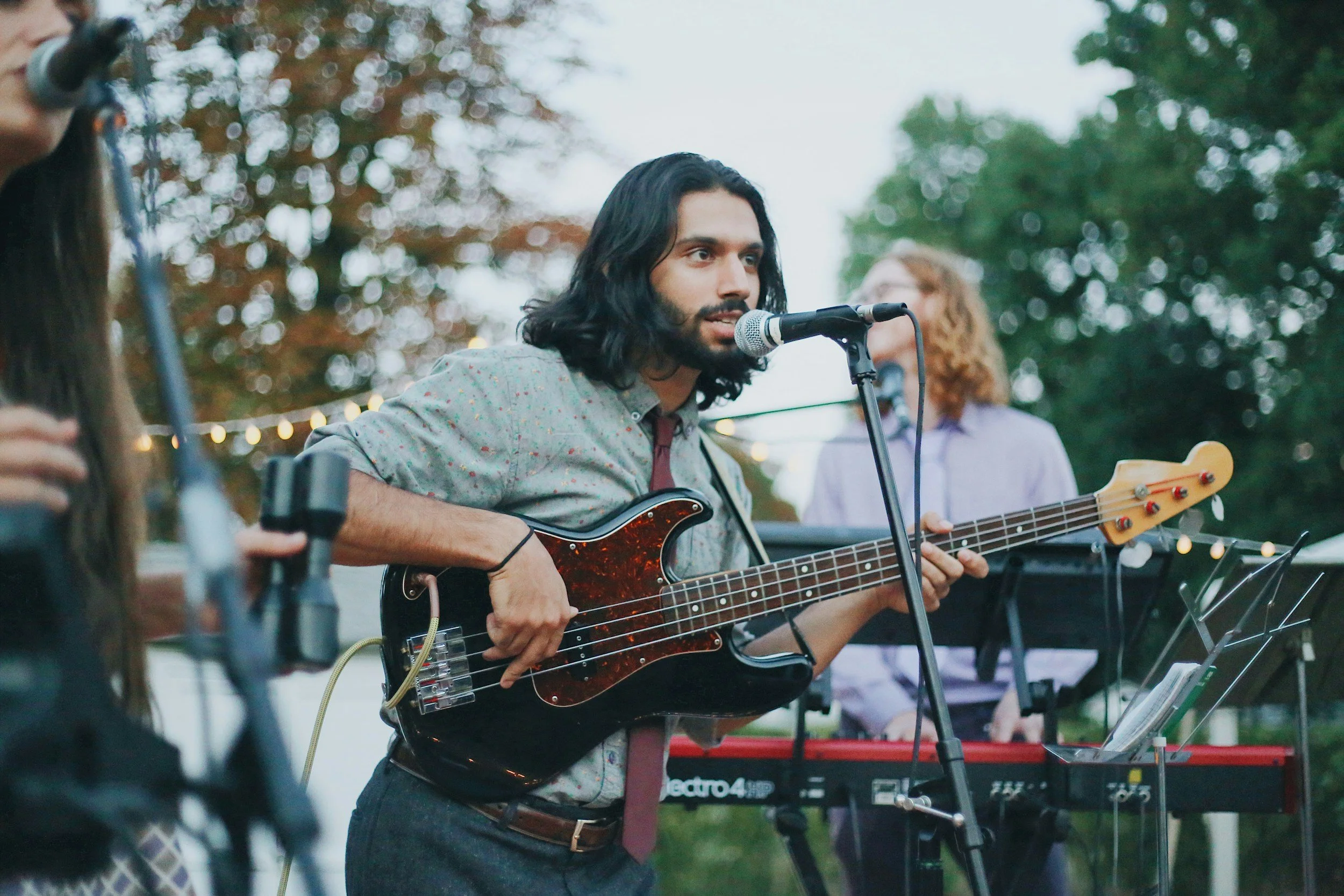 A man with shoulder-length black hair and a beard playing an electric bass guitar at an outdoor music event. He is singing into a microphone, with a keyboard player in the background and string lights in the setting.