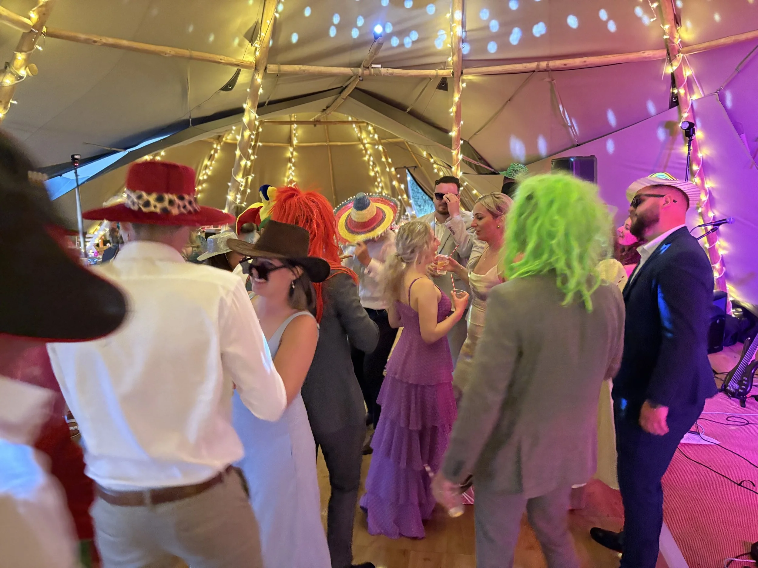 People dancing and socializing at a wedding under a tent decorated with string lights, wearing colorful hats and costumes at a lively party.