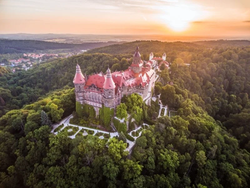 A fairytale-style castle with red roofs and multiple turrets situated on a hill, surrounded by lush green trees during sunset.