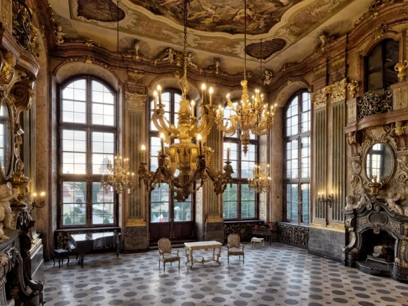 Luxurious European-style room with large arched windows, ornate gold chandeliers, intricate wall moldings, and antique furniture, including chairs and a fireplace.
