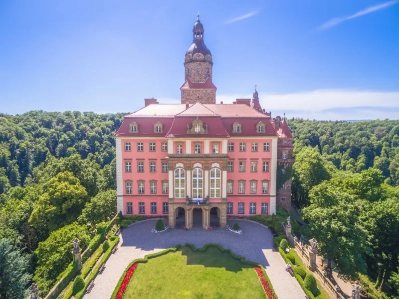 A large pink castle with a tower on top, surrounded by green trees and a landscaped garden in the front.