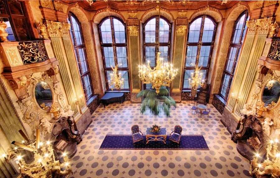 Luxurious grand room with tall, arched windows, ornate gold and cream decor, chandeliers, a grand piano, and elegant furniture.