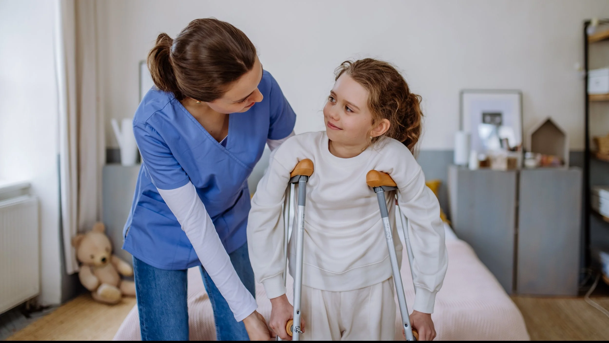 A nurse helping a young girl with leg braces standing on crutches in a bedroom.