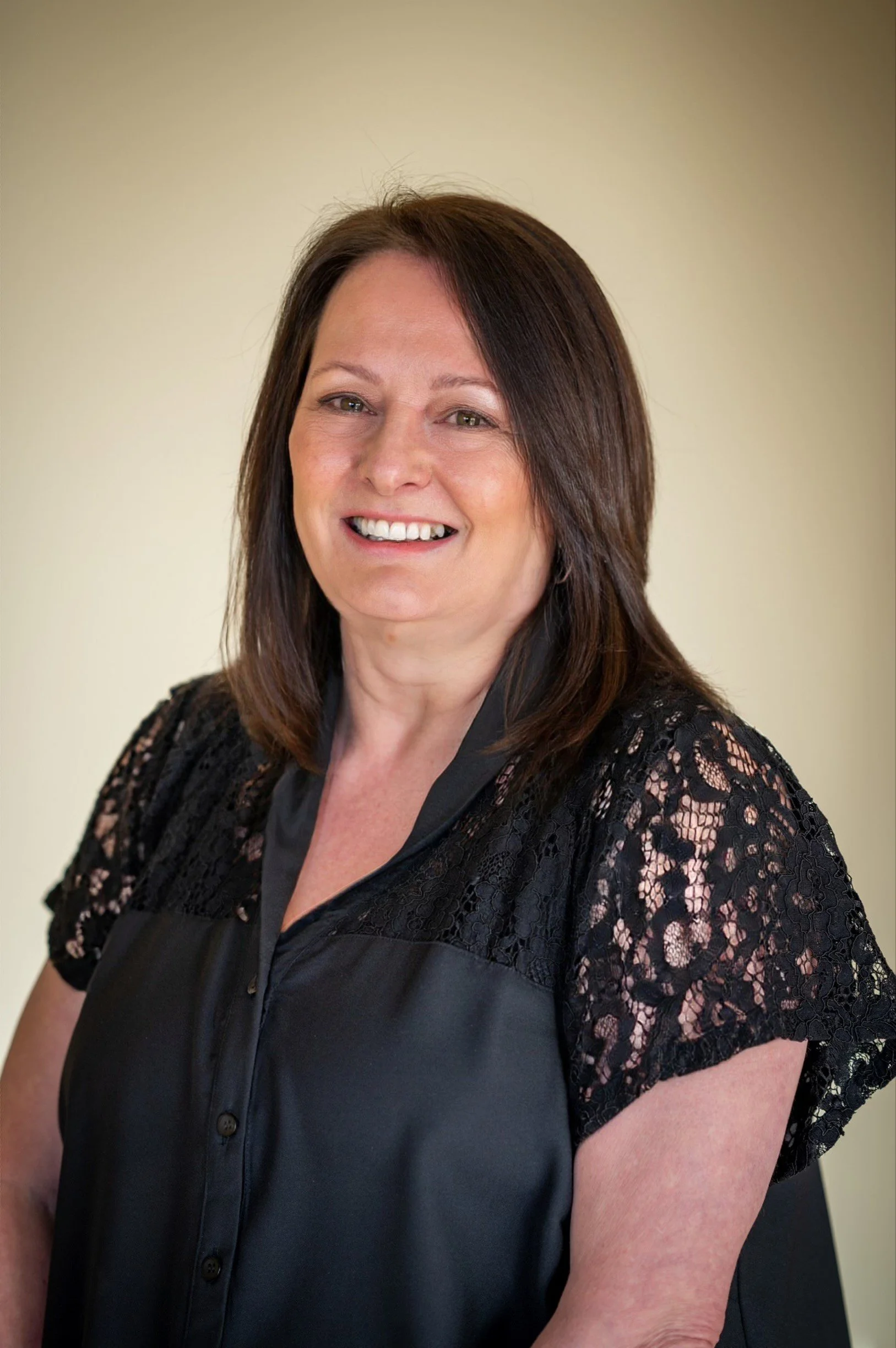 A woman with shoulder-length dark brown hair, smiling, wearing a black satin blouse with lace sleeves, standing against a plain light-colored background.