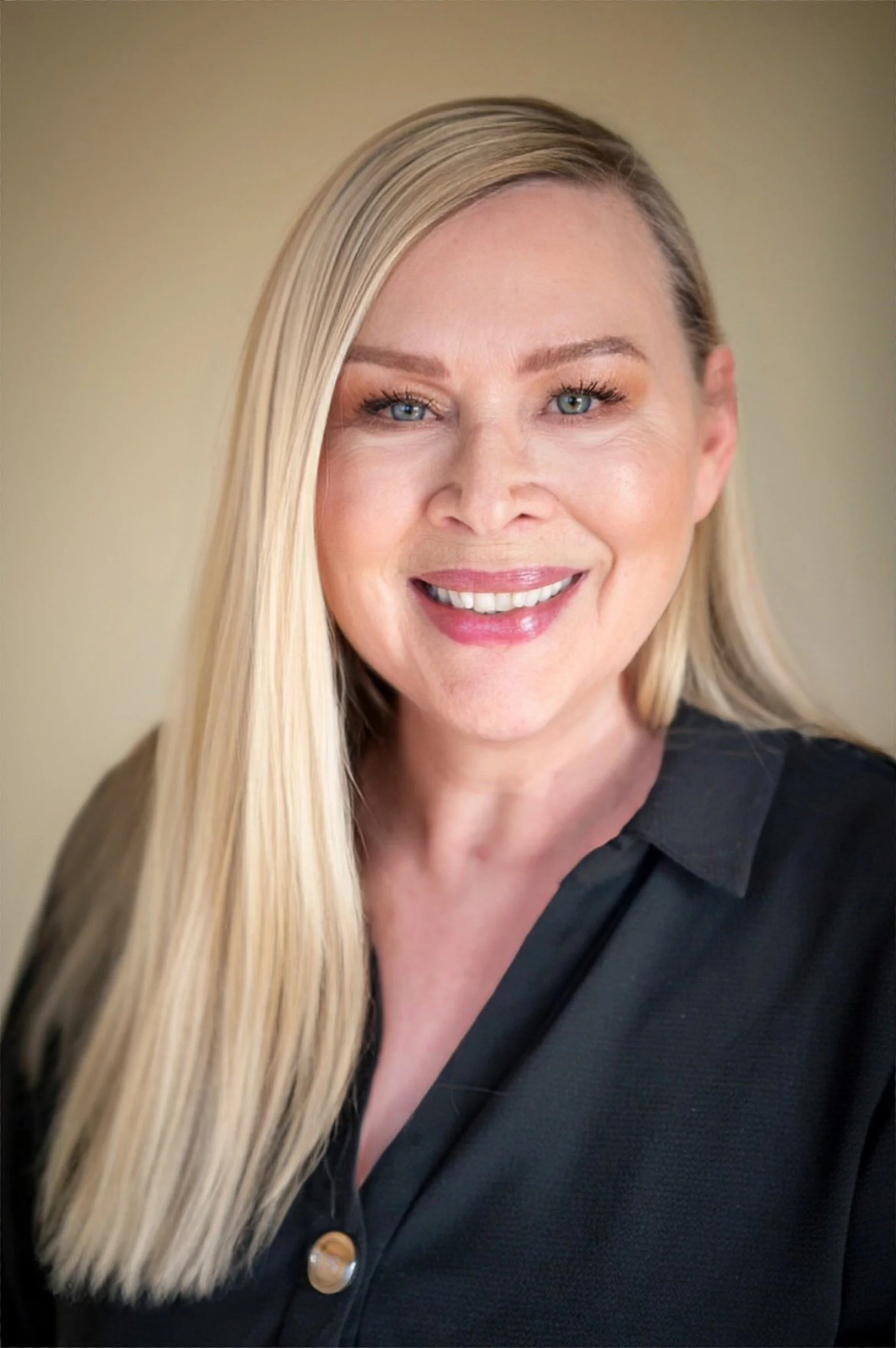 A portrait of a woman with long blonde hair, blue eyes, and fair skin, smiling and wearing a black top with a pearl button