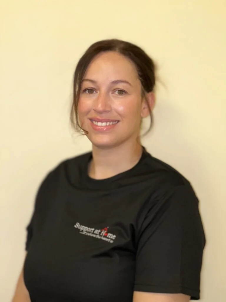 A woman with brown hair tied back, smiling, wearing a black t-shirt with a logo that says support at home, standing against a beige background.