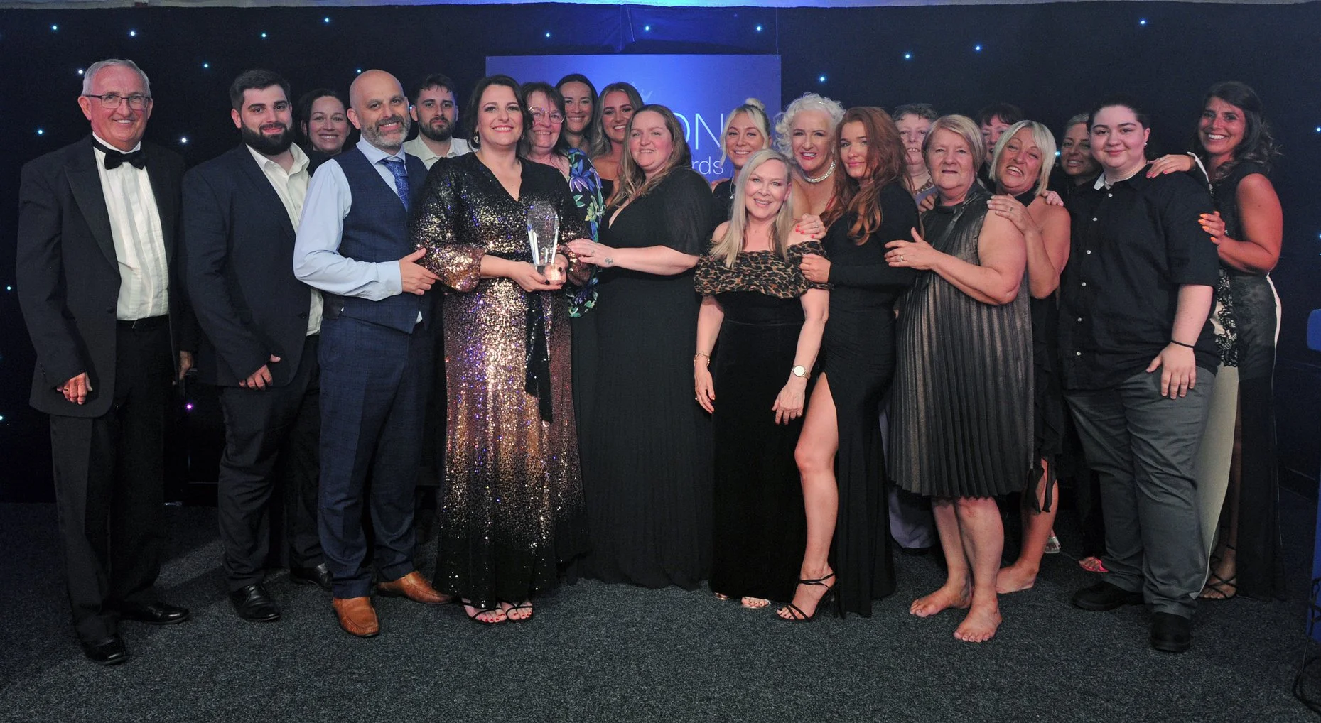 Group of men and women in formal attire at awards ceremony, one woman holding an award, standing in front of a backdrop with stage lights.