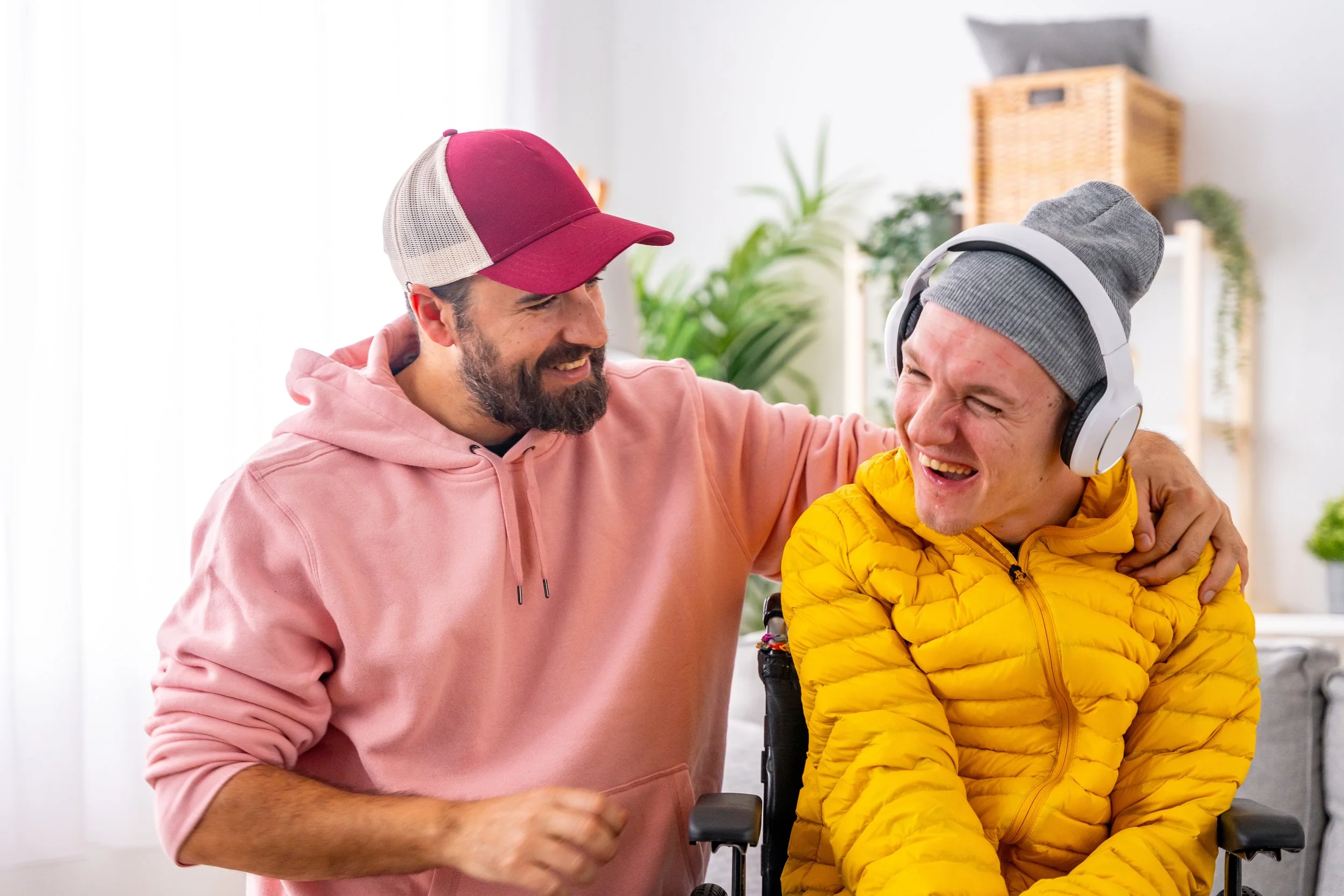 Two men smiling, one in a pink hoodie and cap, the other in a yellow jacket and gray beanie, sharing a joyful moment indoors.