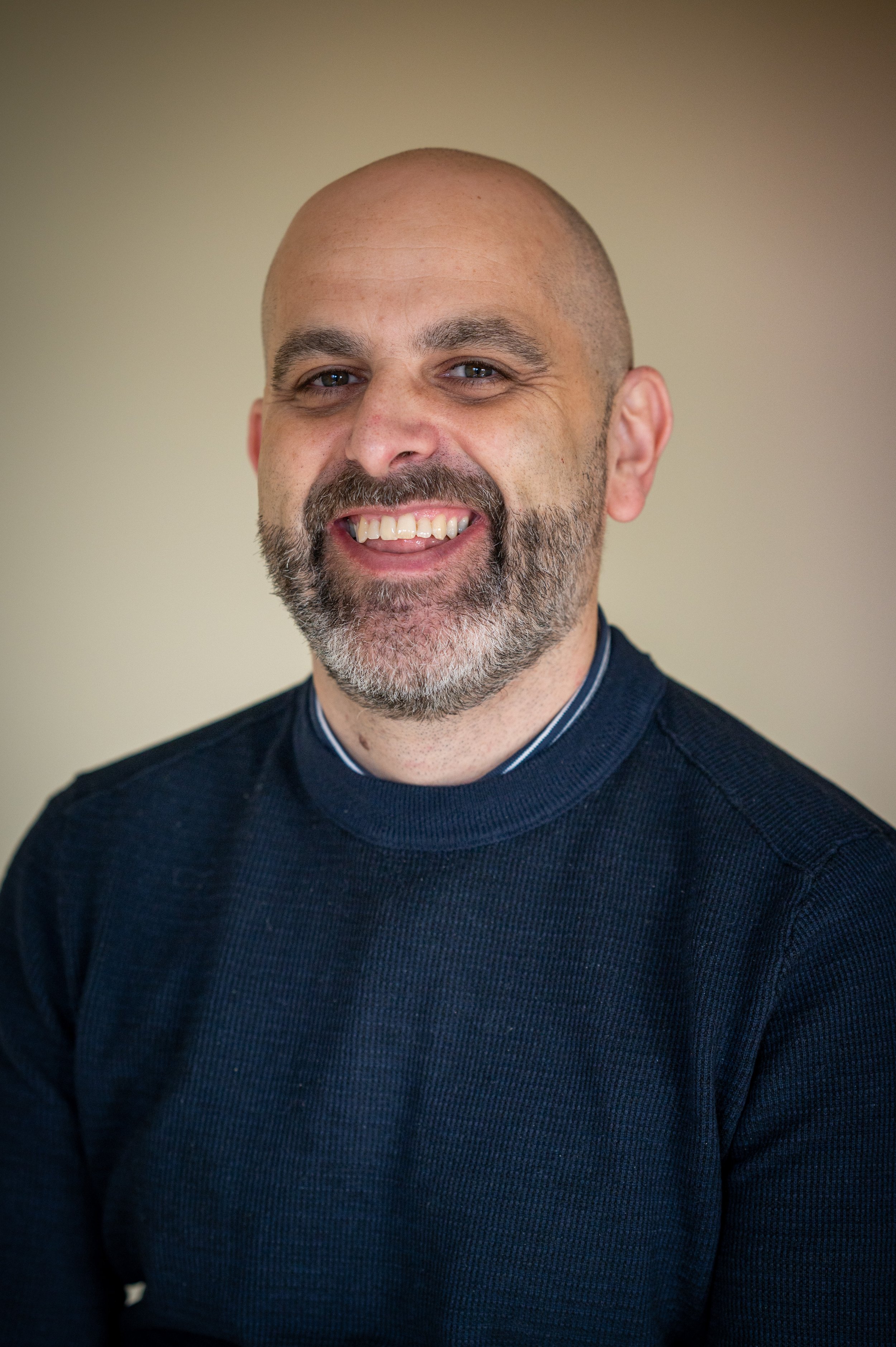 A smiling bald man with a beard, wearing a dark blue sweater, against a neutral background.
