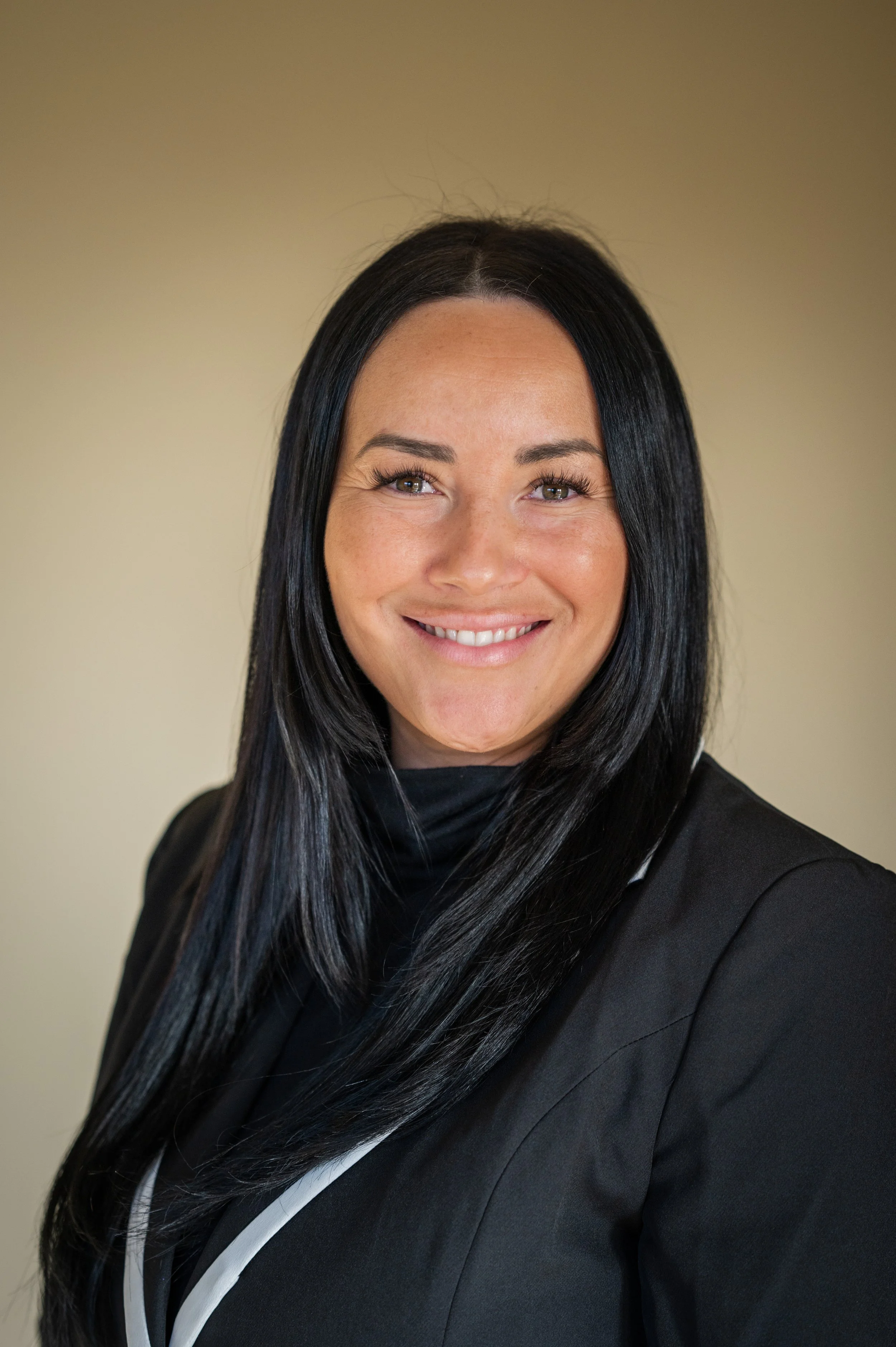 Professional woman with long black hair smiling at the camera, wearing a black blazer and black top, against a beige background.