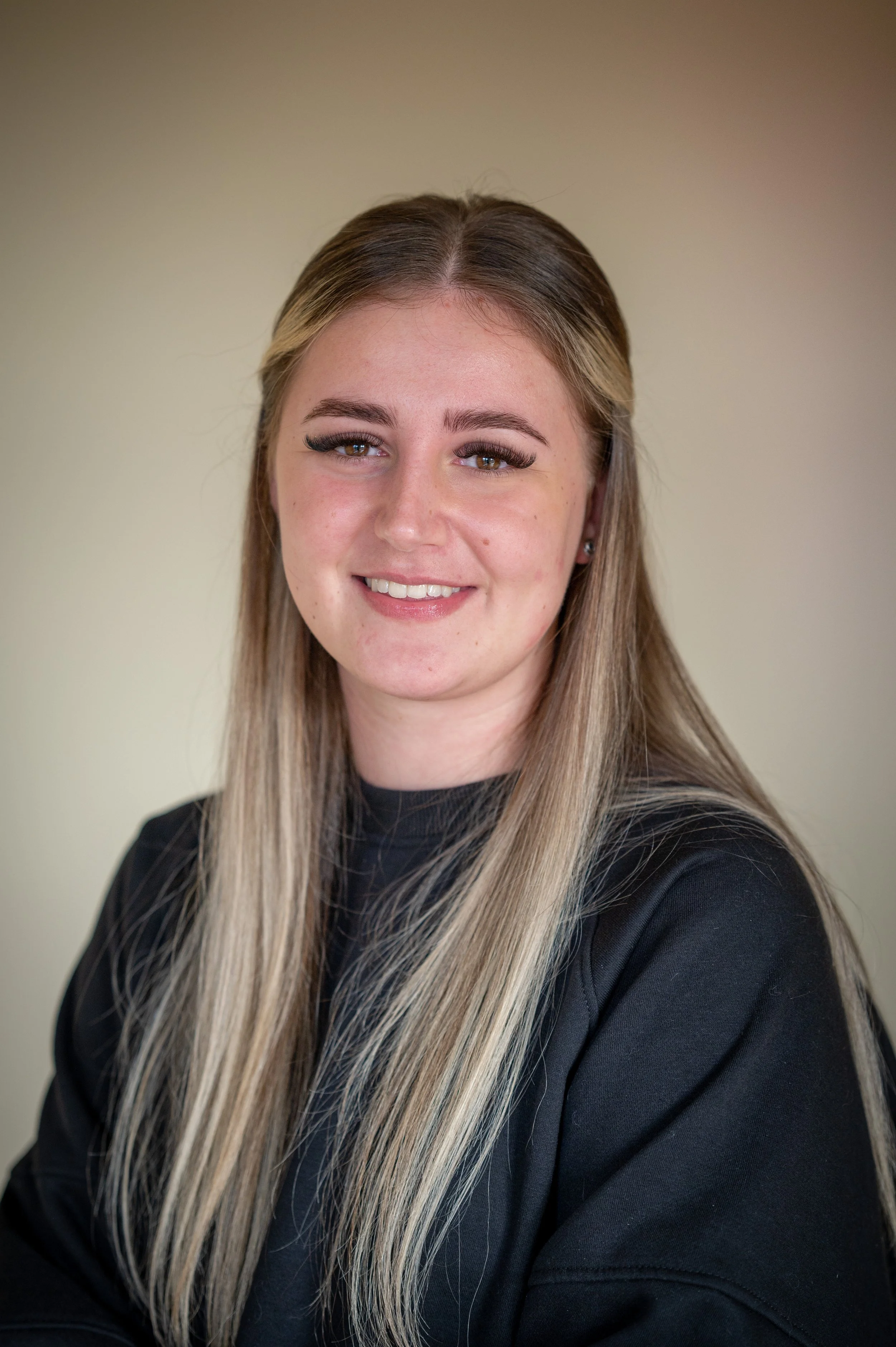 A smiling young woman with long blonde hair and brown eyes, wearing a black top, sitting against a neutral background.