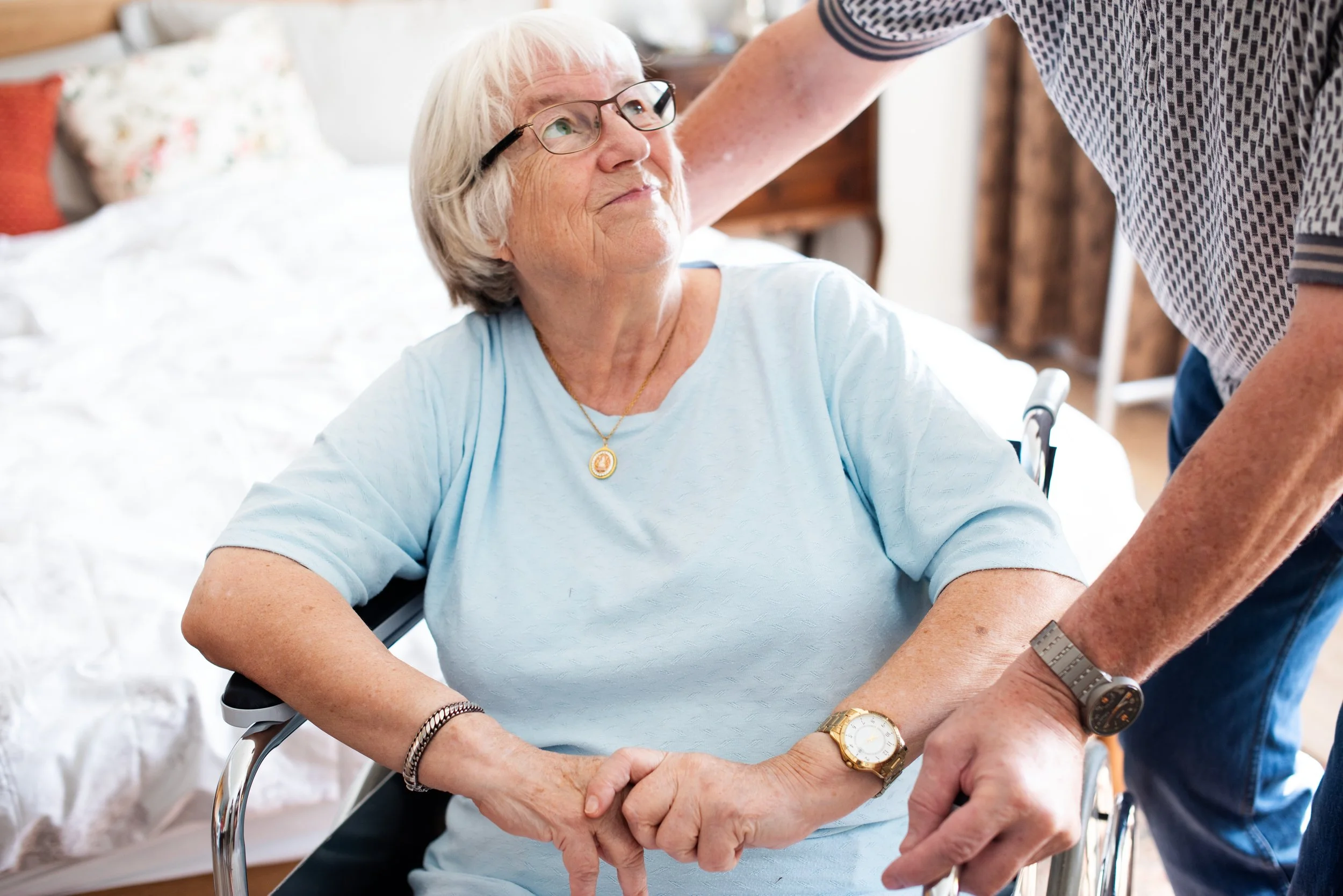 An elderly woman with glasses sitting in a wheelchair holding hands with a man who is helping her.