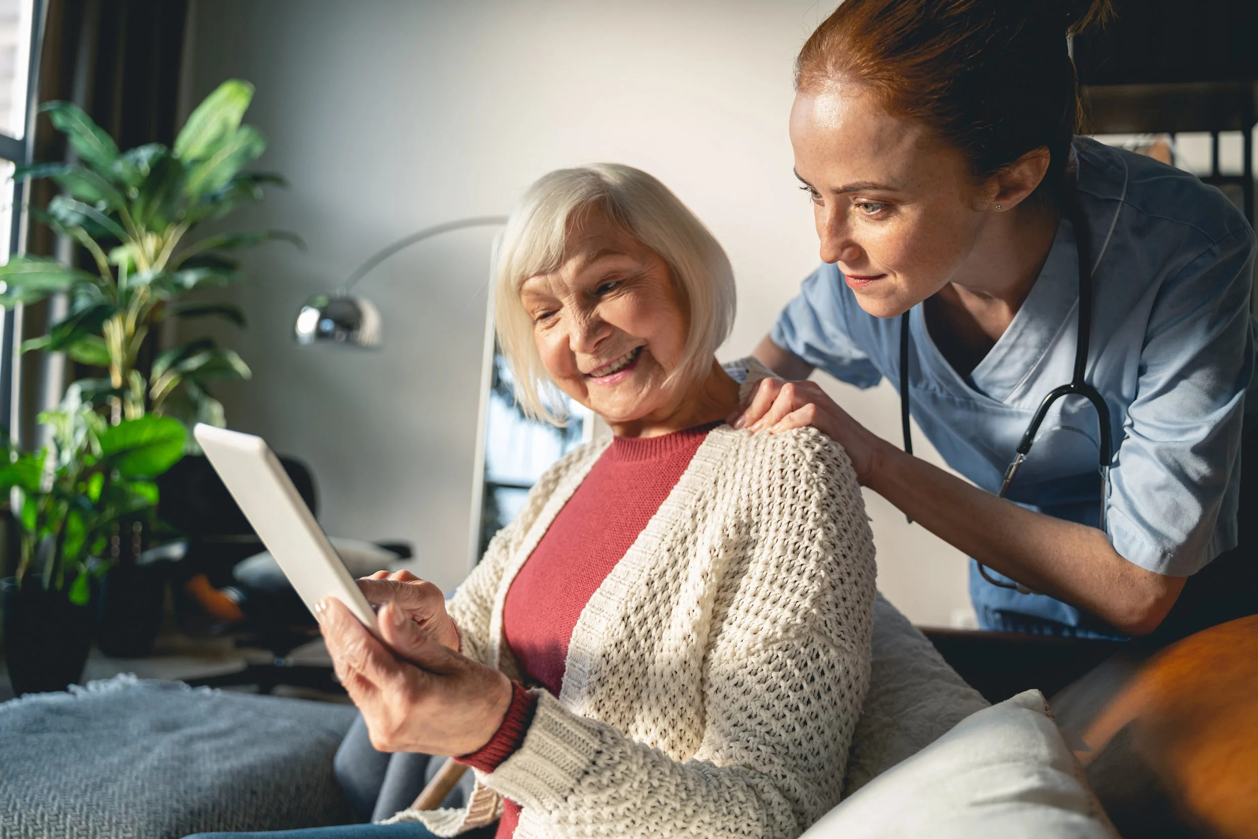 A nurse interacting with an elderly woman who is sitting on a couch, smiling, while holding a tablet. The scene takes place in a cozy, well-lit room.