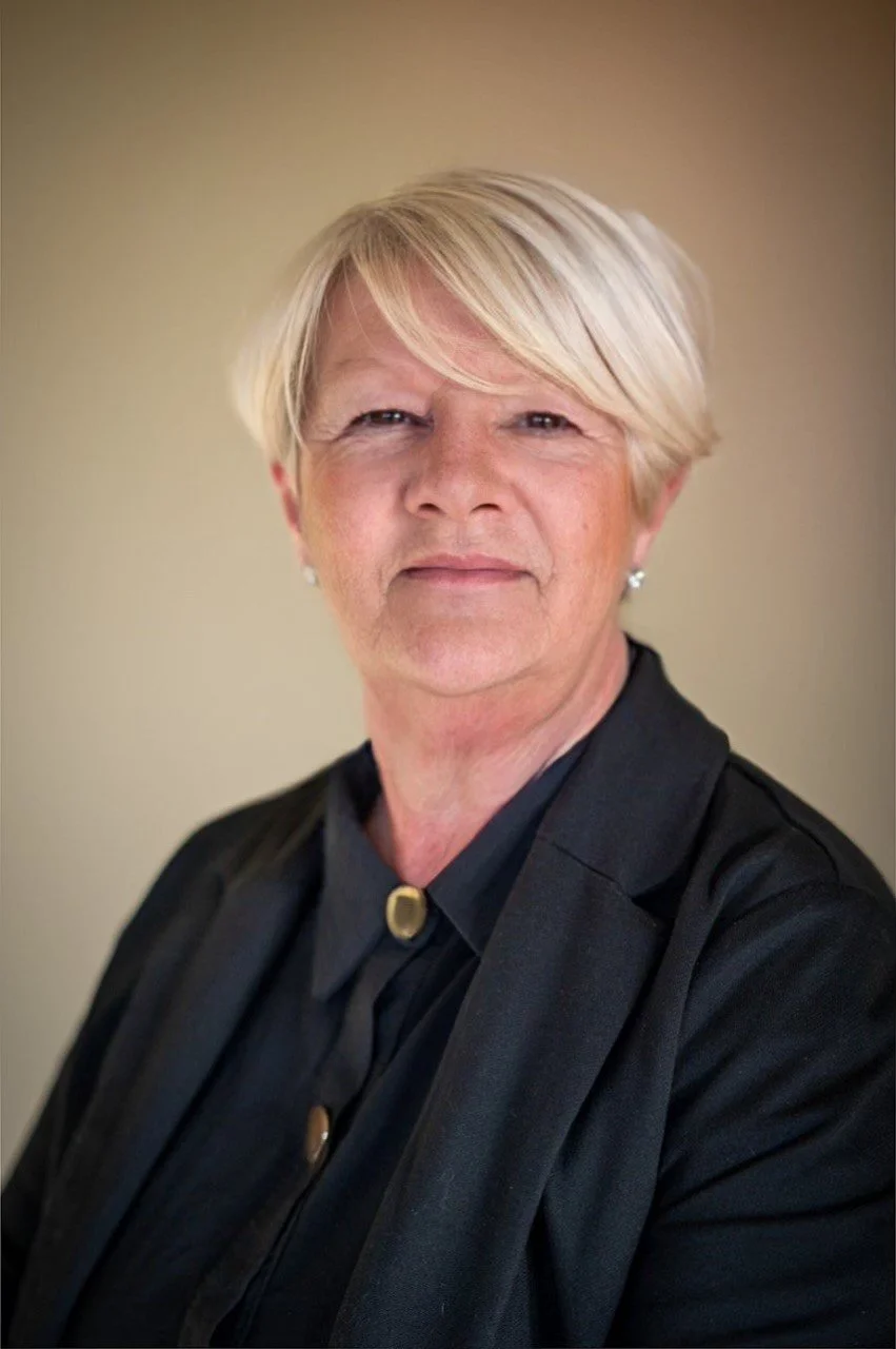 A professional woman with short blonde hair, wearing pearl earrings and a black blazer over a dark shirt, is posing against a plain background.