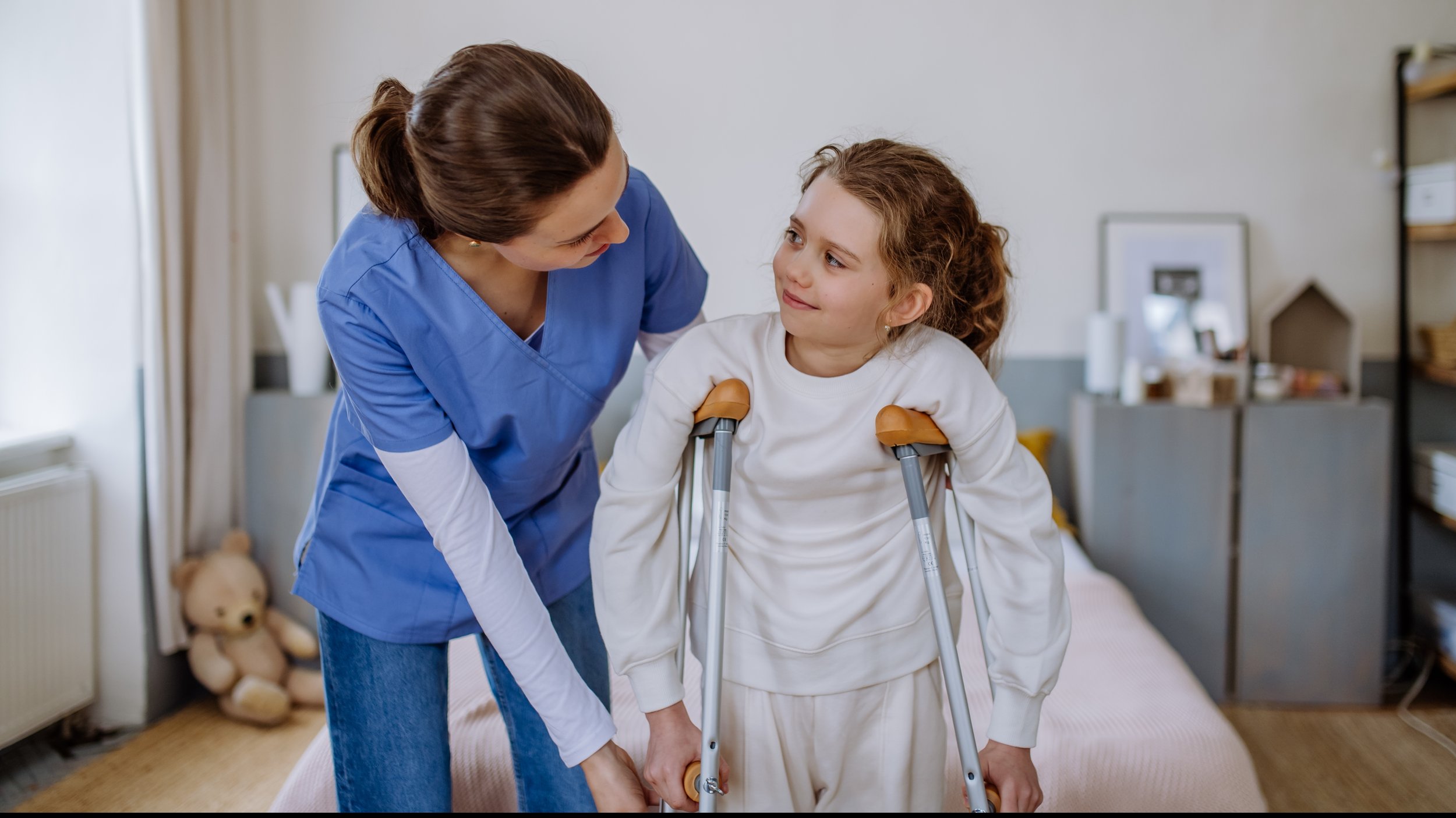 A nurse assisting a young girl with crutches in a room with a bed, a teddy bear, and shelves.