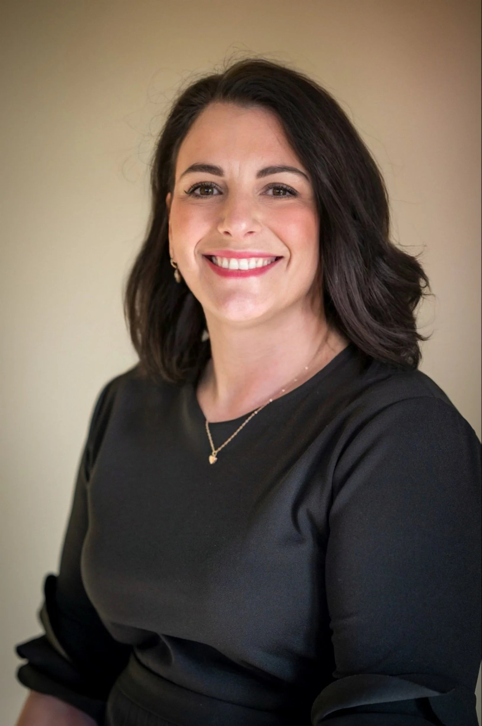 A woman with dark brown hair, wearing a black top with three-quarter sleeves, a necklace, and earrings, smiling in front of a plain beige wall.