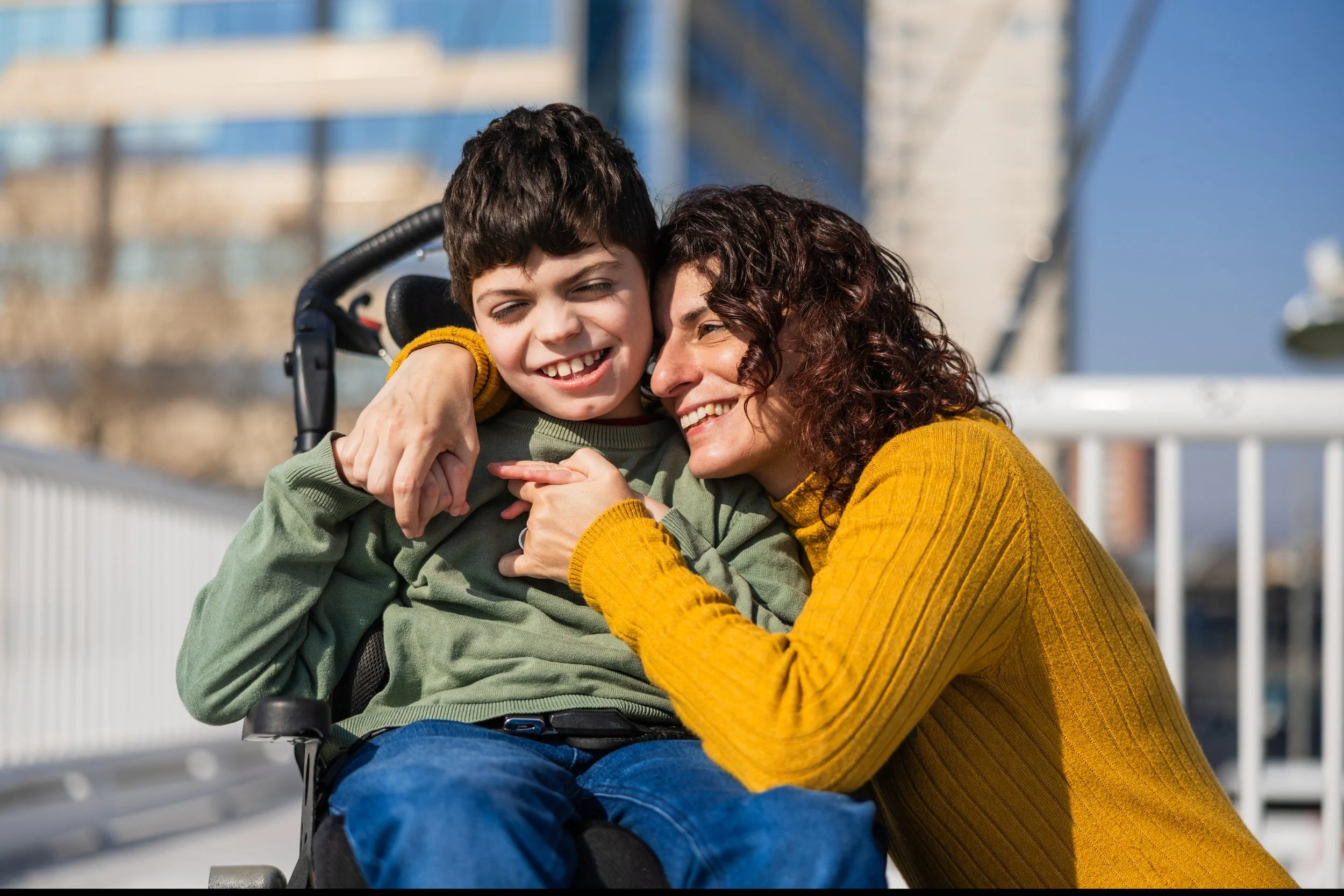 A smiling woman embracing a young boy in a wheelchair outdoors with a bridge and blue sky in the background.