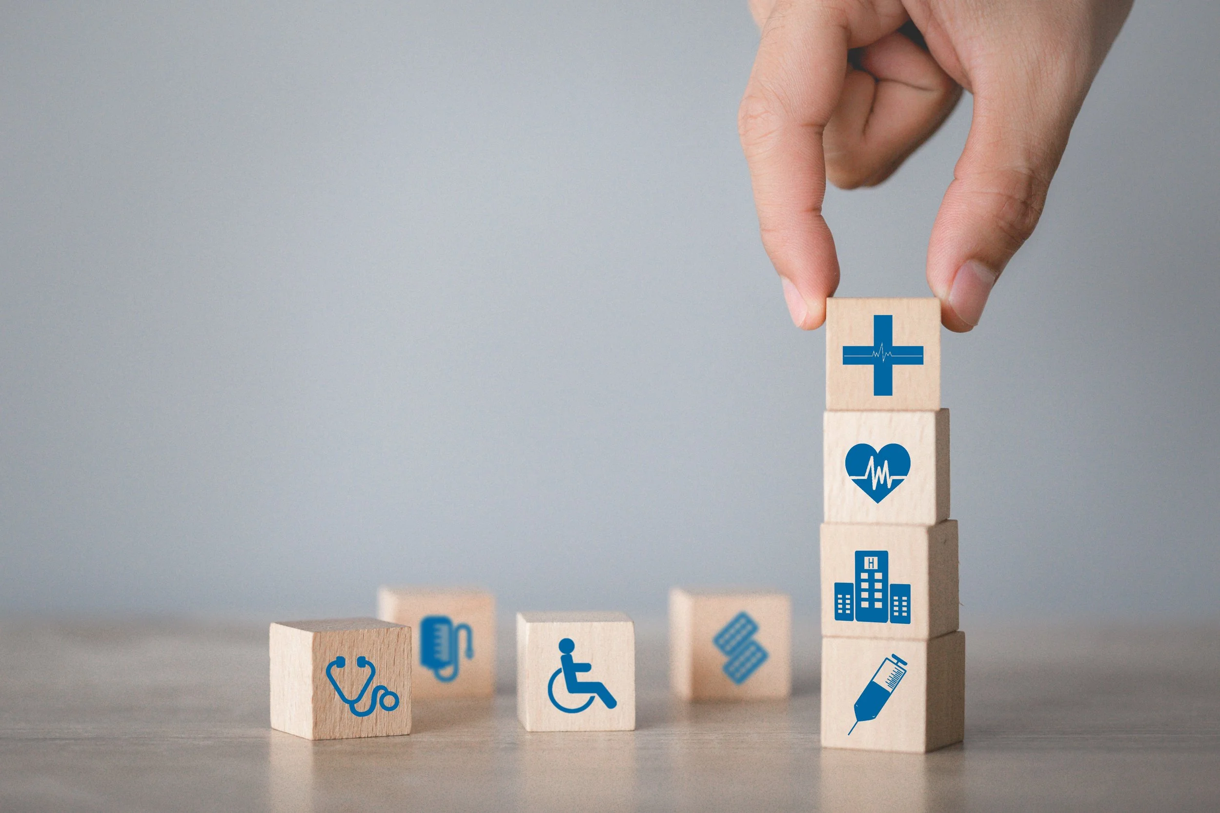 A hand stacking wooden blocks with medical icons, including a cross, heart, hospital, and syringe, on a wooden surface with additional medical-related blocks laying down.